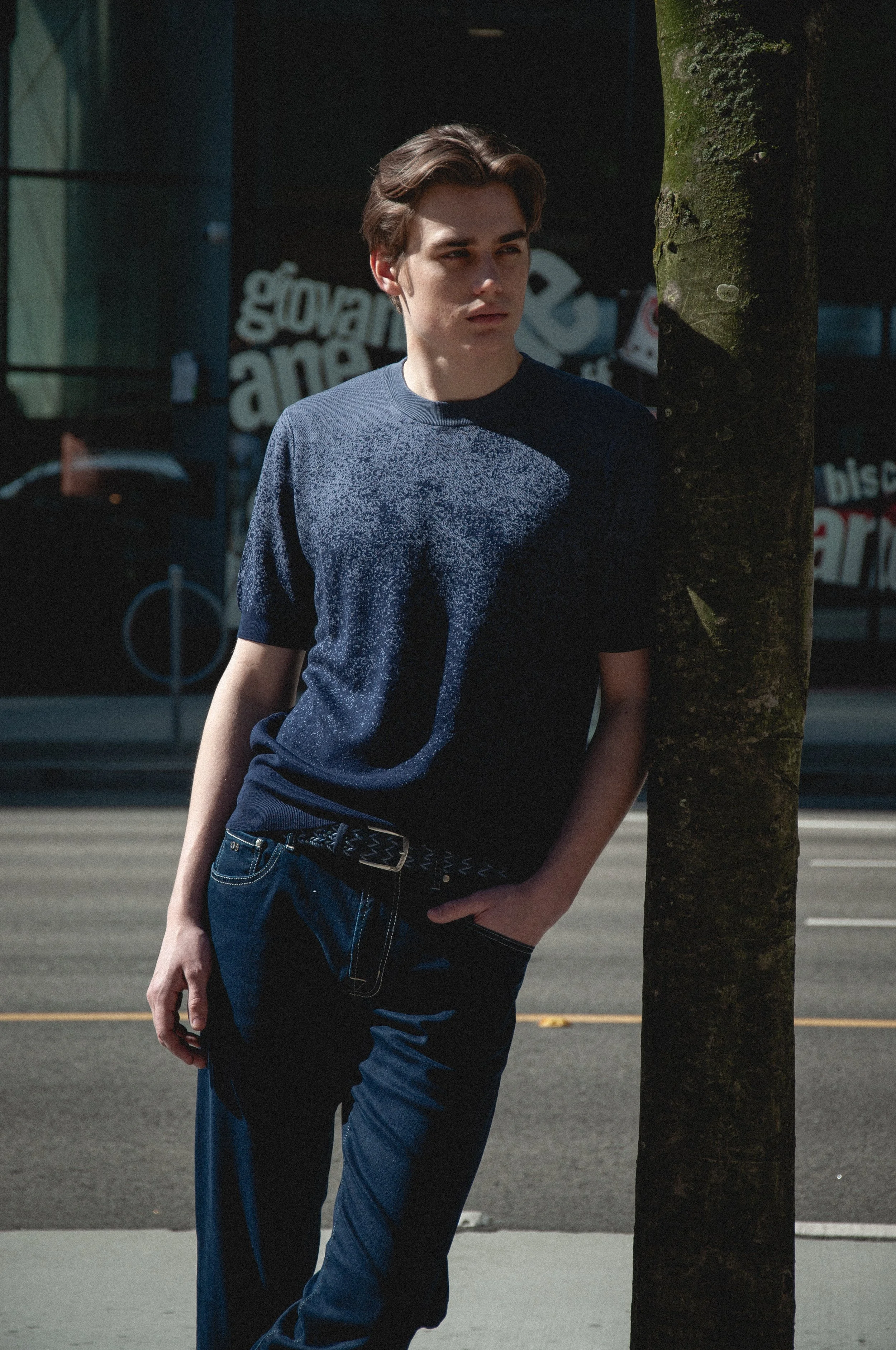 Young man with brown hair standing outside next to a tree, wearing a blue shirt and jeans, with a background of a storefront and a bicycle.