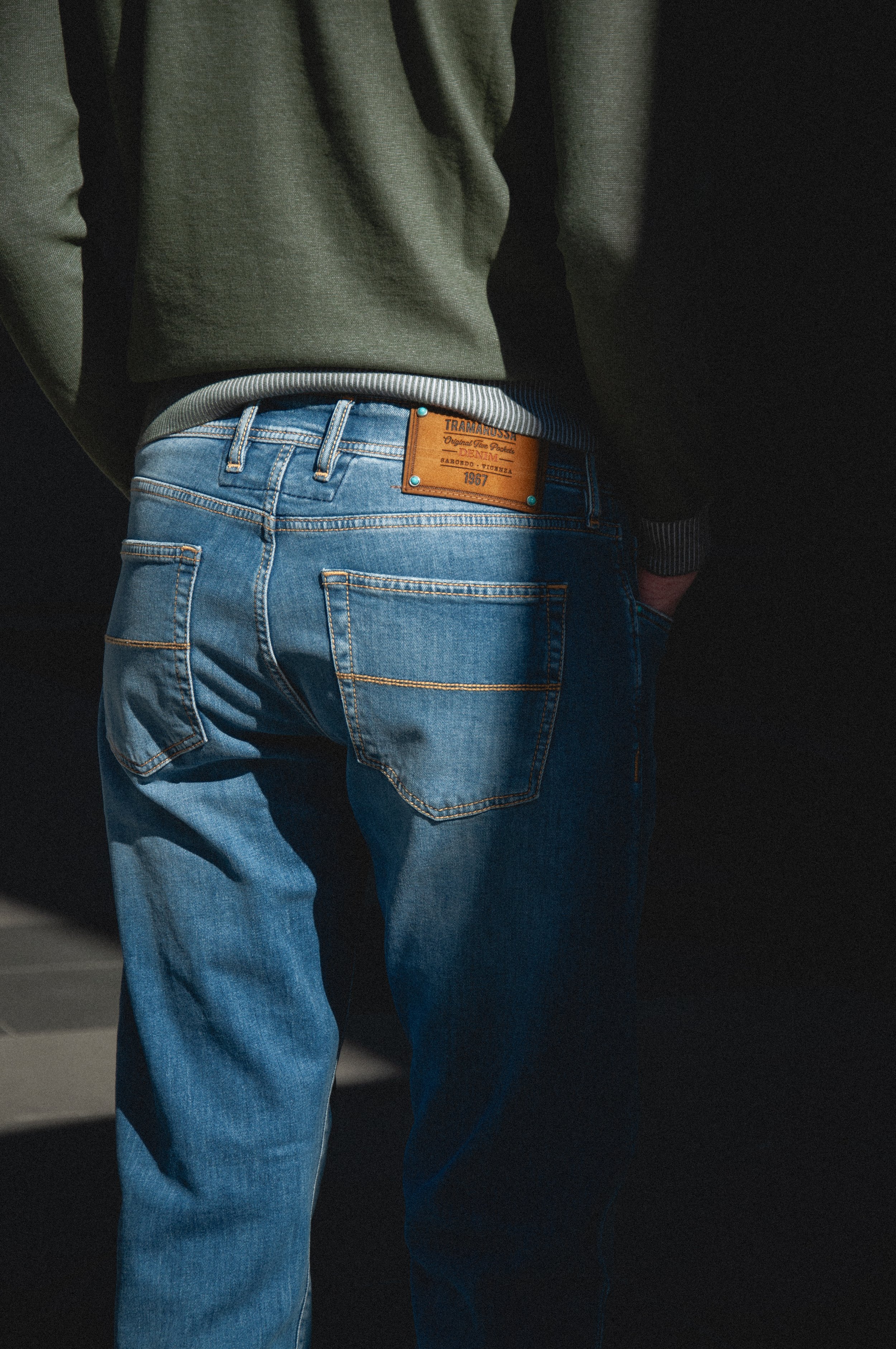 Close-up of a person's lower back and hips, wearing a green sweater, blue jeans, and a brown leather patch on the waistband of the jeans, with a dark background.