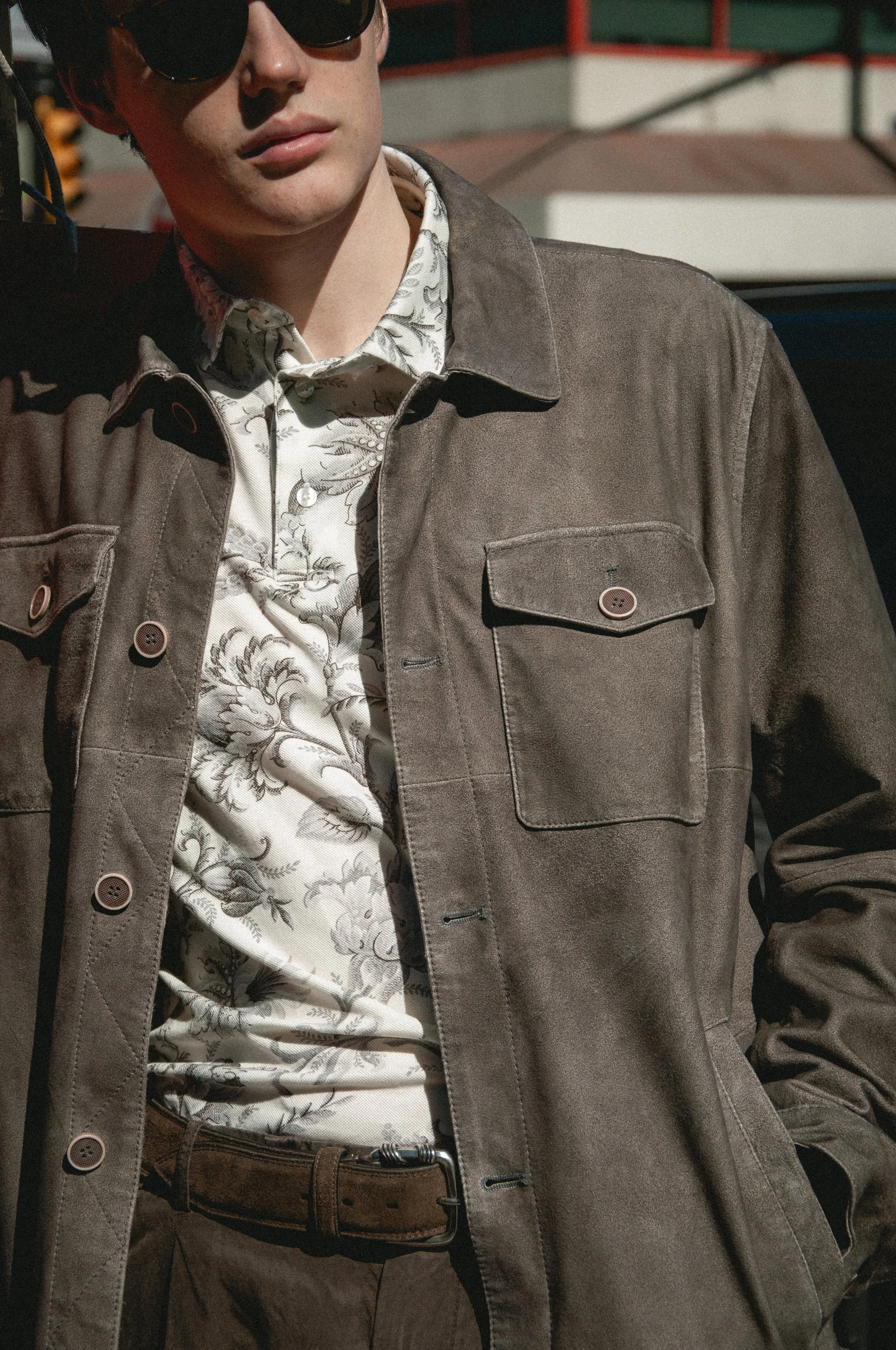 Close-up of a person with dark hair wearing sunglasses, a patterned shirt, a brown jacket, and a brown belt, standing outdoors in bright sunlight.