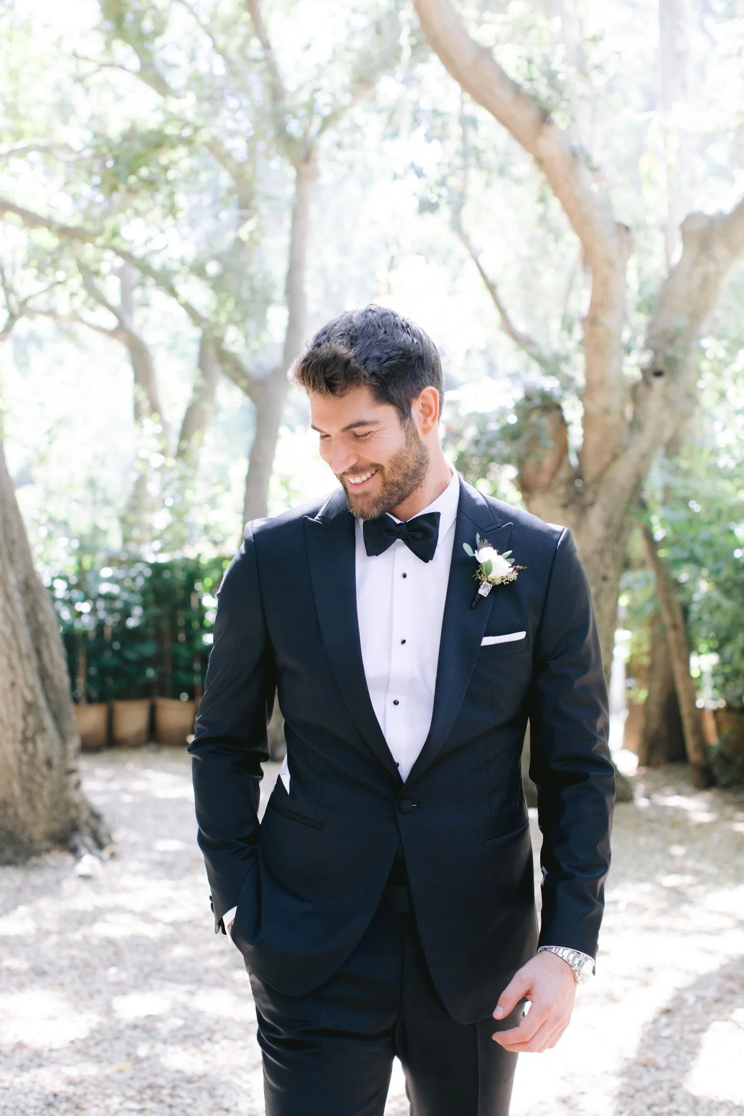 A man dressed in a black tuxedo with a bow tie, smiling and looking down, standing outdoors among trees with sunlight filtering through.