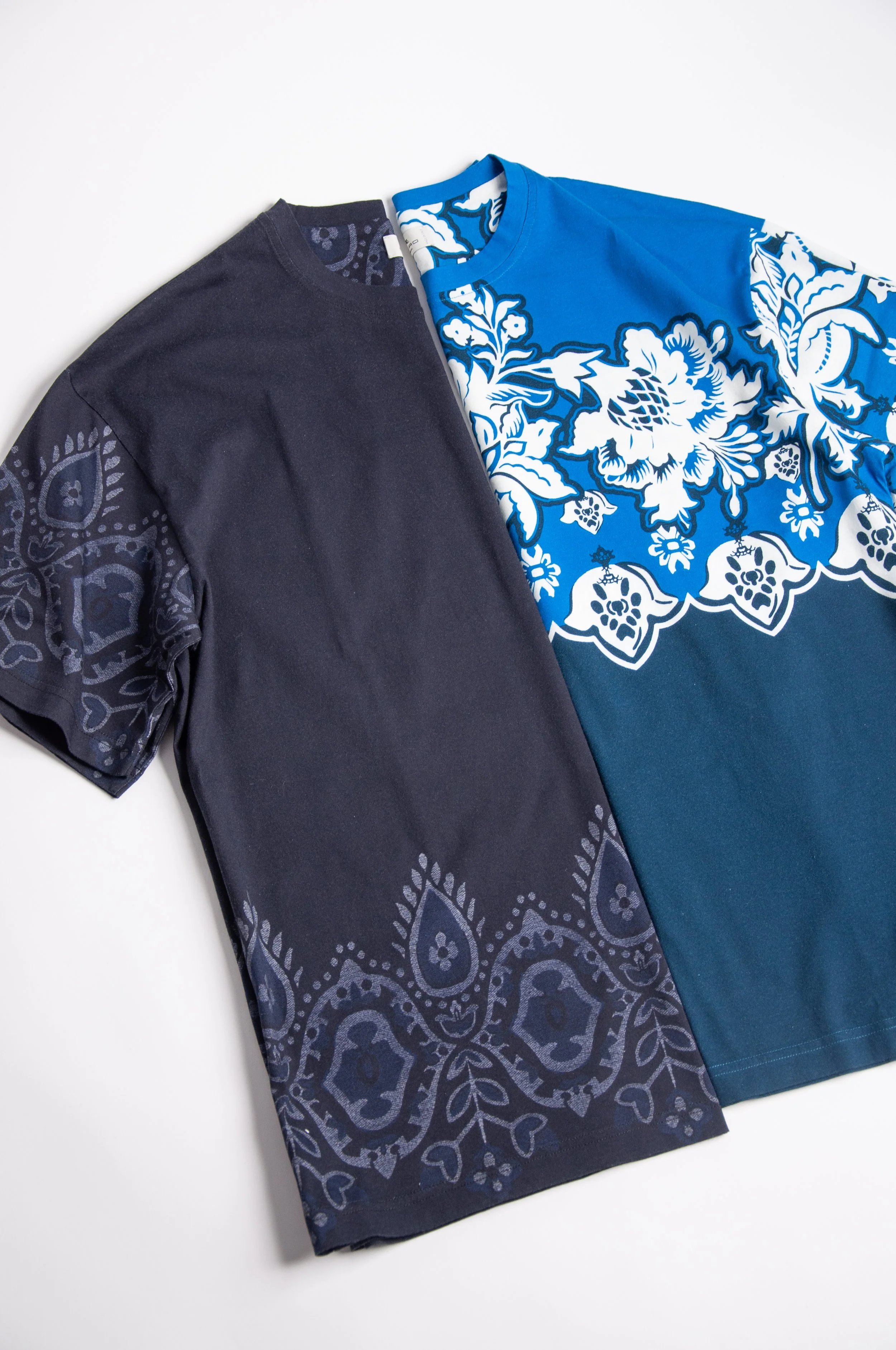 Two T-shirts with bandana-like paisley patterns, one in black with dark pattern and the other in bright blue with white pattern, laid flat on a white surface.