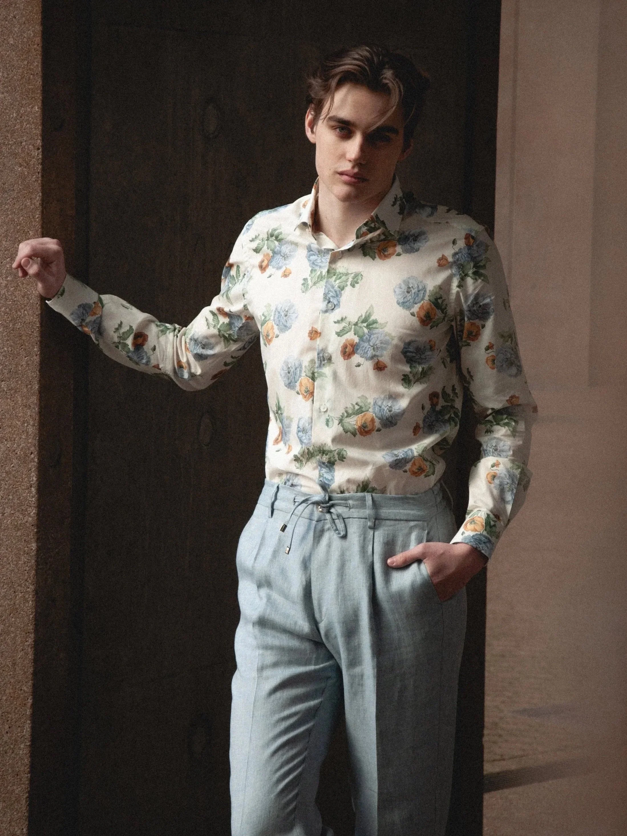 Young man with dark hair, wearing a floral patterned button-up shirt and light gray pants, standing indoors with one hand in his pocket and the other resting on a wall.