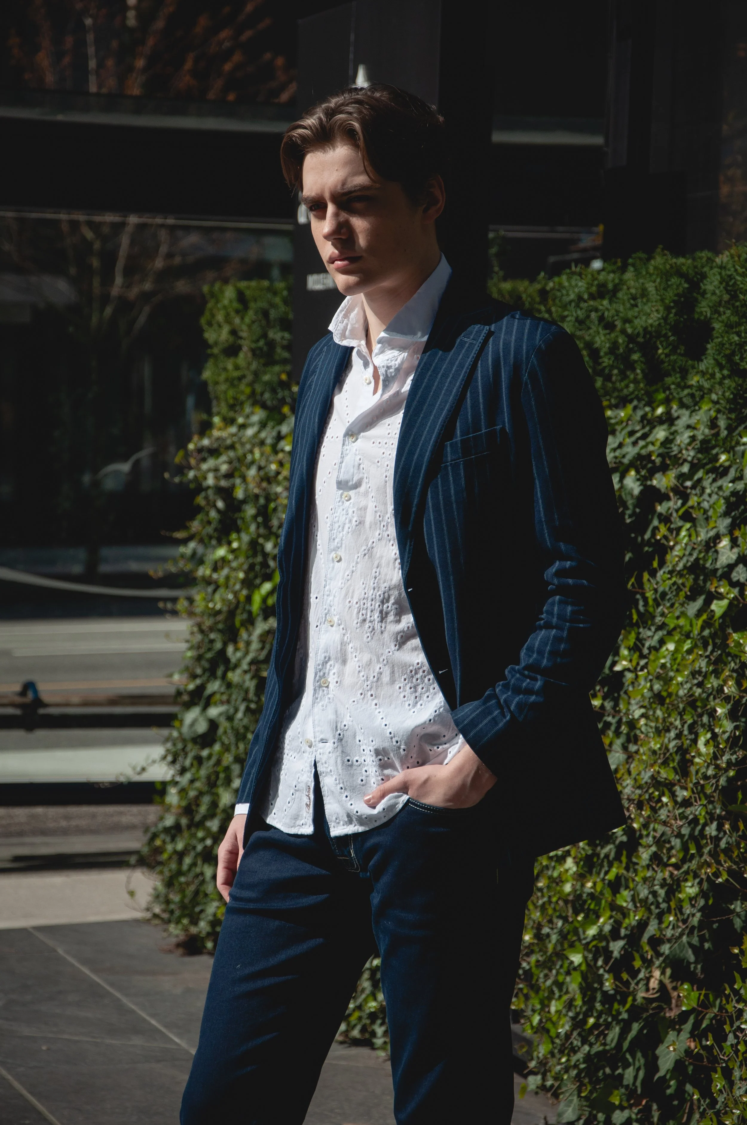 A young man in a dark pinstripe blazer and white embroidered shirt stands outdoors, with one hand in his pocket, looking thoughtfully to the side with sunlight highlighting his face and hair, against a background of green bushes and a modern building.