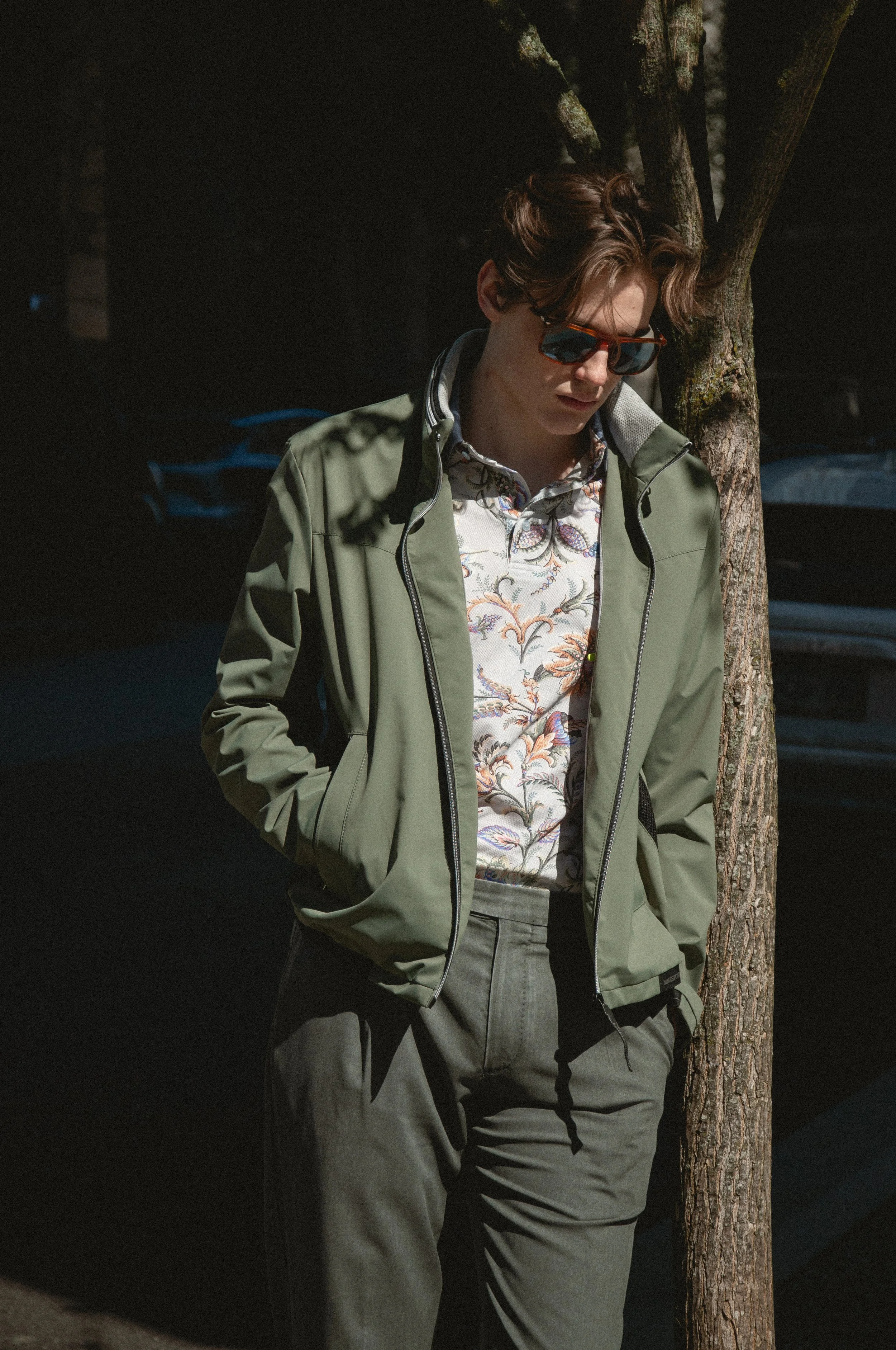 A young man with brown hair wearing sunglasses, a floral shirt, a green jacket, and gray pants leans against a tree in a shaded outdoor area.