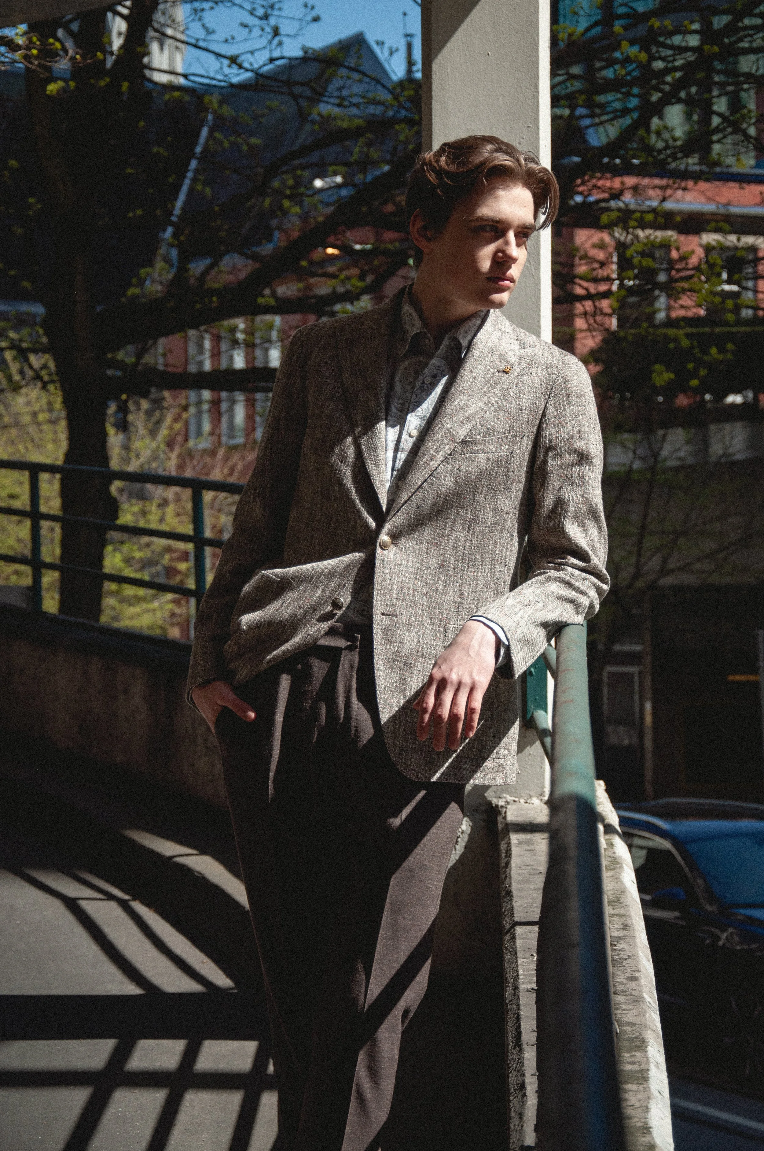 A young man with brown hair wearing a beige blazer and dark pants leaning against a white pole outdoors in daylight, with trees and buildings in the background.