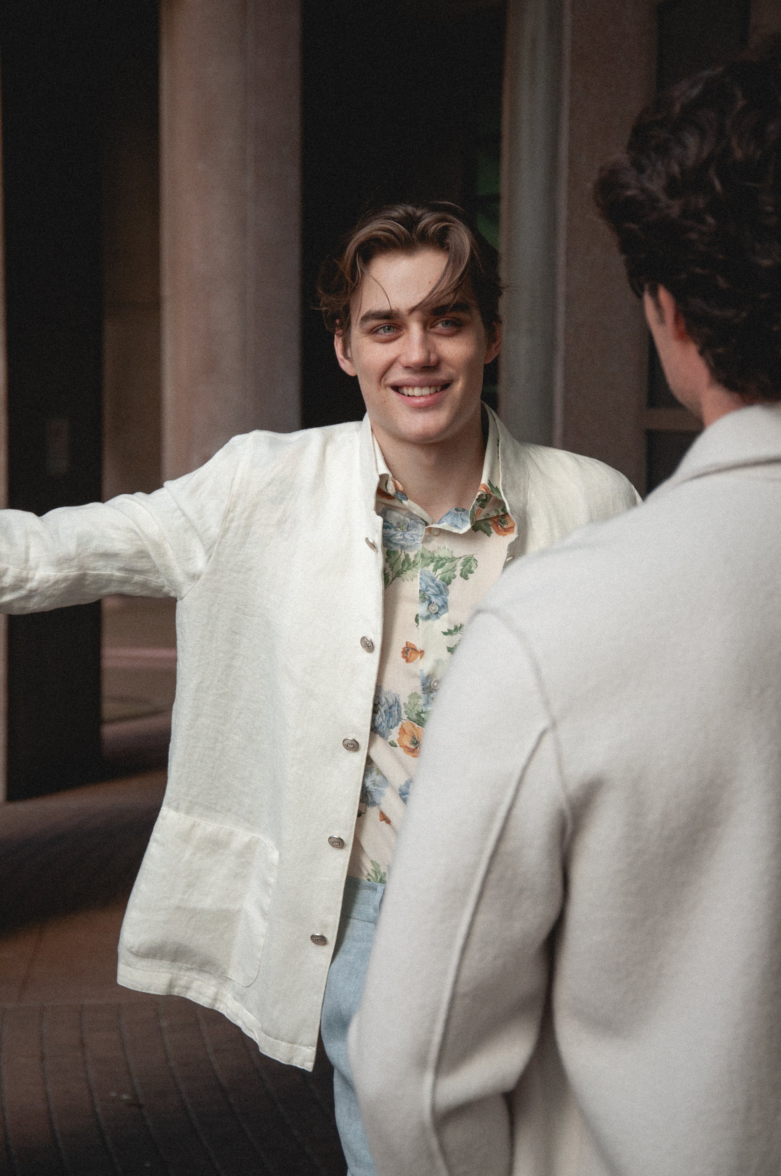 Two men in conversation, one smiling, the other with curly hair, both wearing light-colored suits, standing indoors near a doorway.