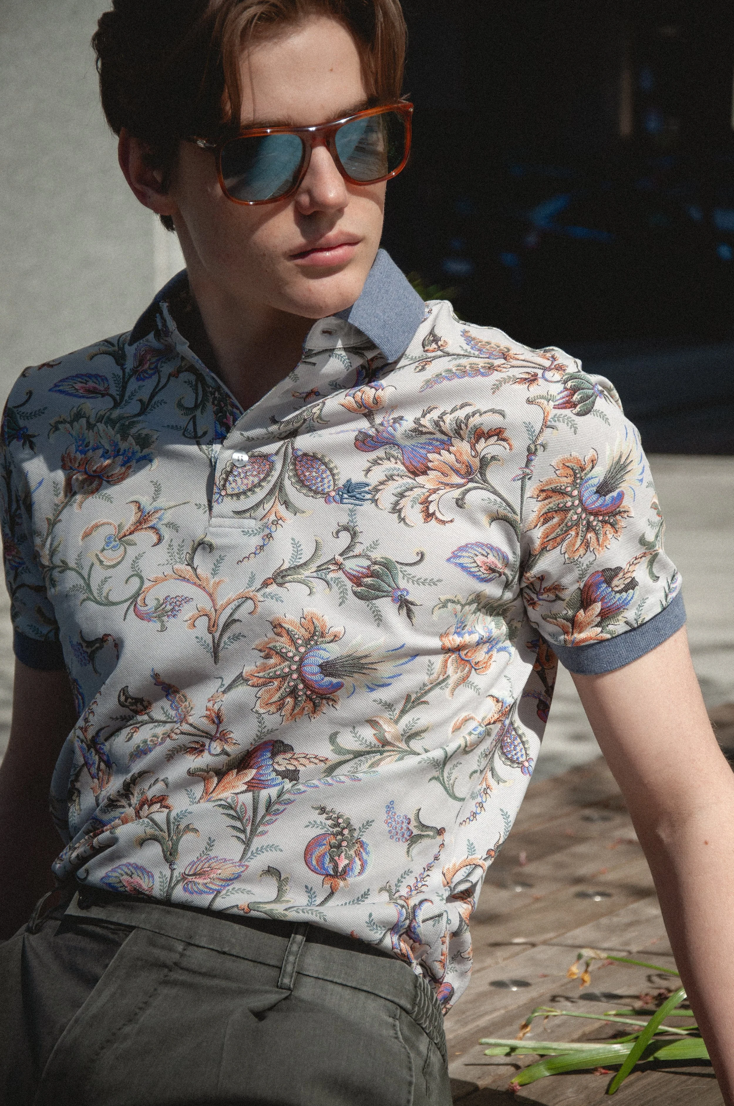 A person with short hair wearing reflective sunglasses, a colorful floral patterned shirt with a denim collar, and gray pants, outside on a sunny day.