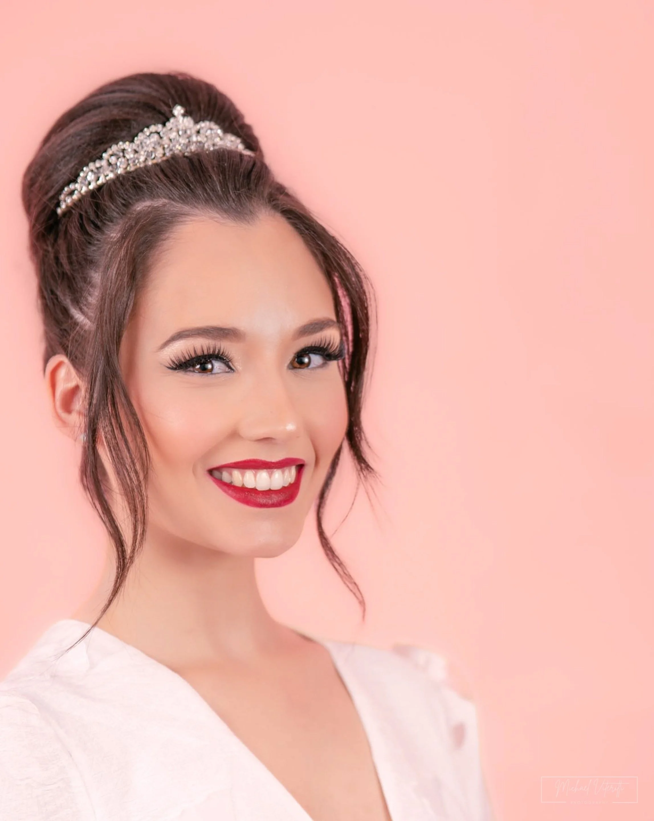 Primo piano di una donna sorridente con trucco impeccabile, capelli raccolti decorati con una tiara di pietre preziose, sfondo rosa