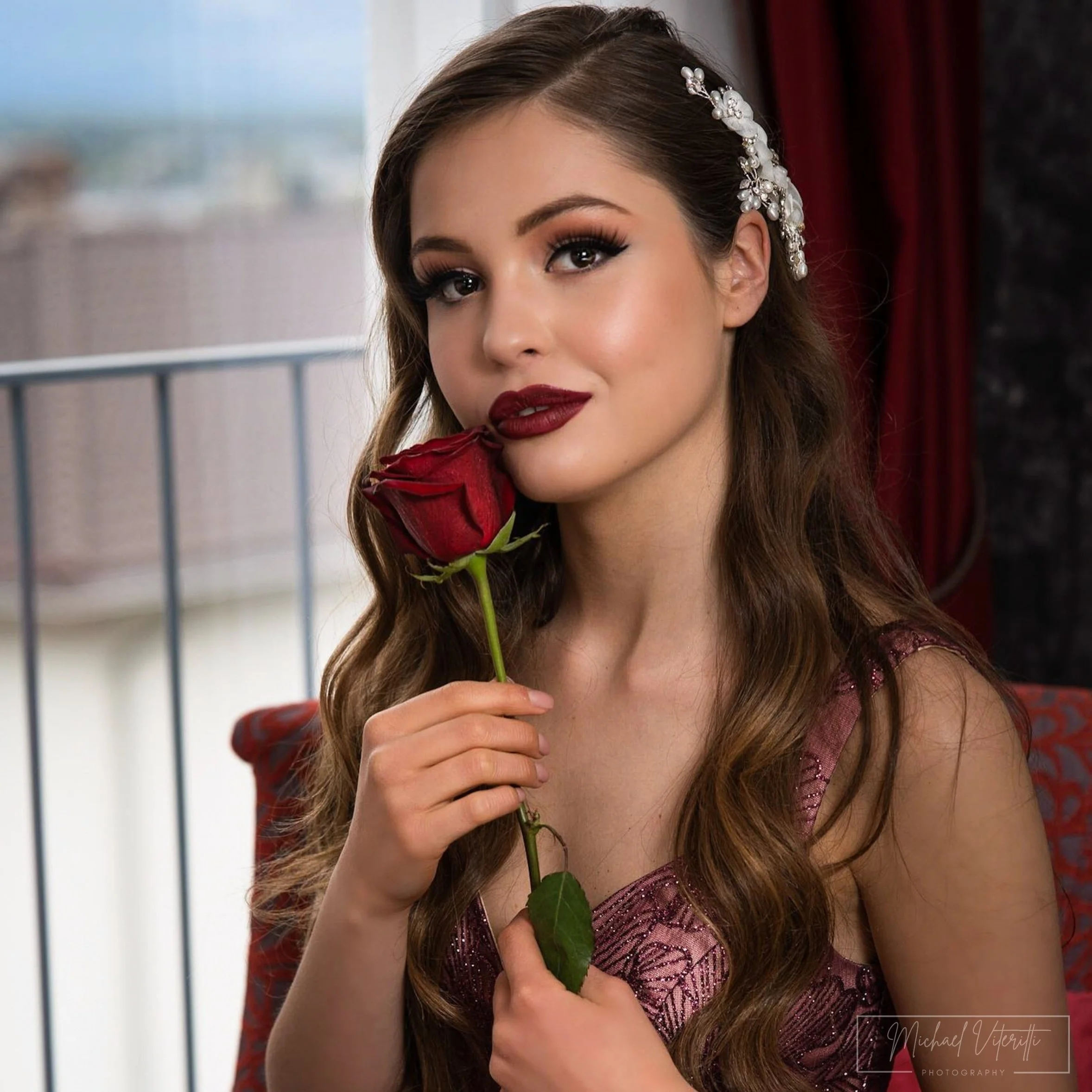 Ragazza con trucco elegante e capelli acconciati, tiene un centro di rosa rossa vicino al viso, indossa un vestito rosa con dettagli lucidi, sfondo con tende rosse e muro nero.