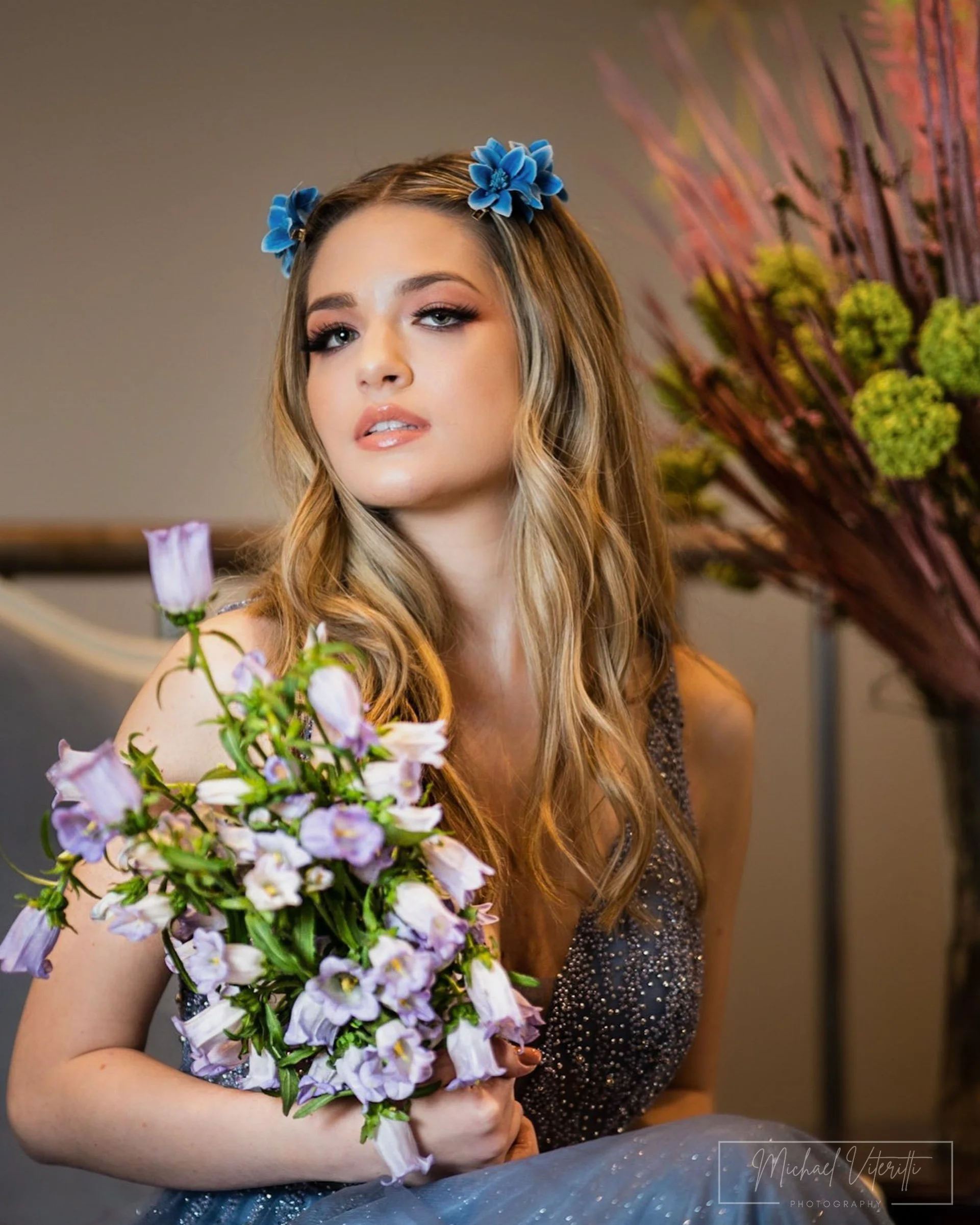 Una giovane donna con lunghi capelli biondi ondulati indossa due fermagli con fiore blu e tiene un mazzo di fiori lilla e ametista. Sullo sfondo si vede un grande ramo decorativo con fiori color verde e rami rossi. La donna indossa un vestito nero co