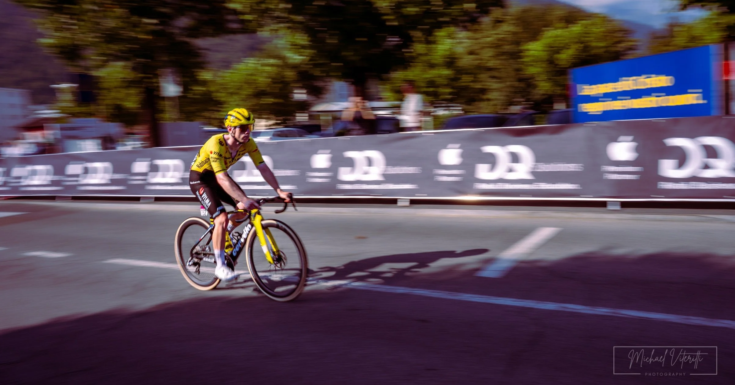 Un ciclista in maglia gialla che corre su una strada di città con sfondo di alberi e cartelloni pubblicitari. L'immagine mostra il movimento rapido del ciclista.