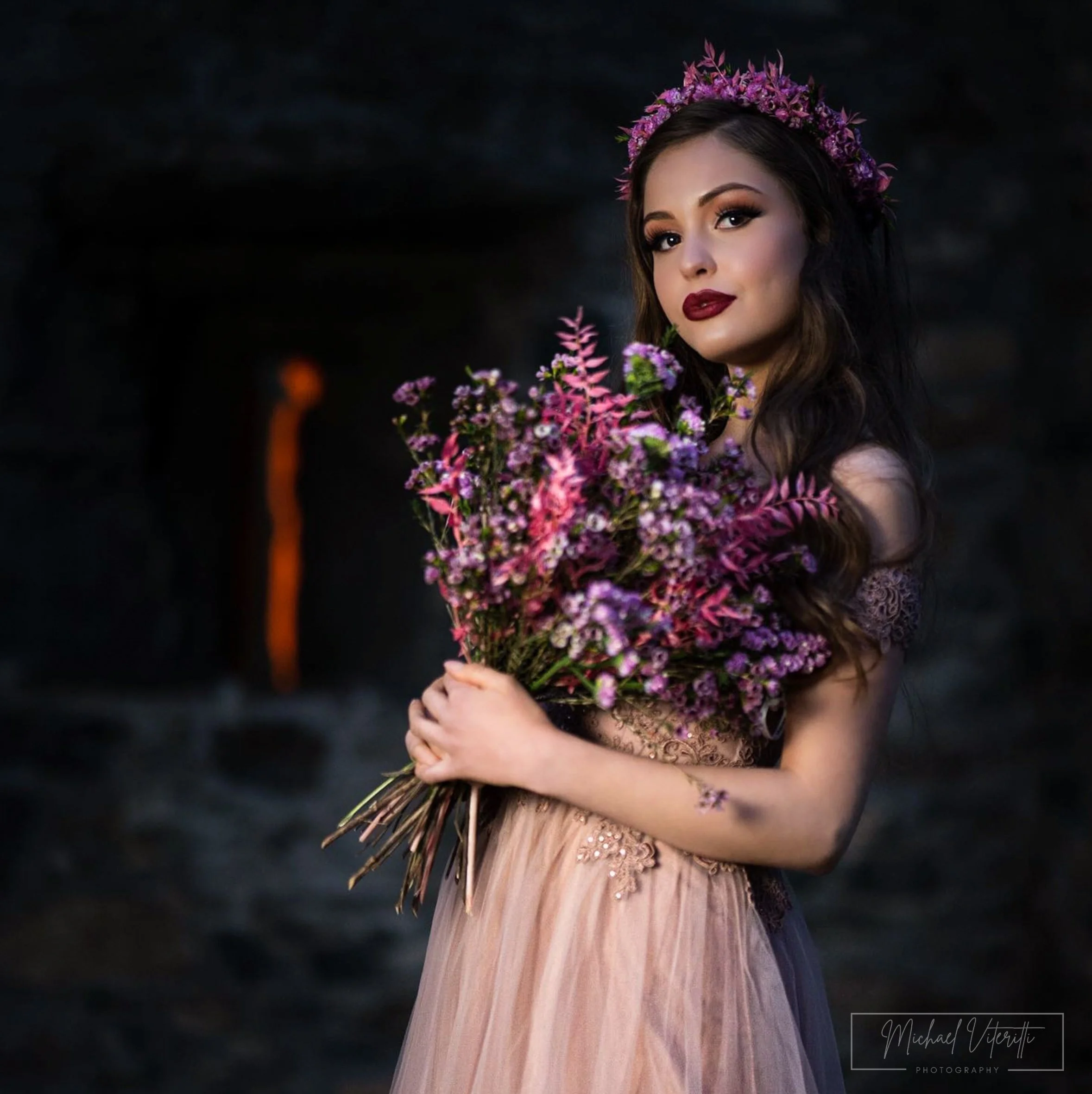 Una donna indossa un vestito di tulle rosa e festeggia con un bouquet di fiori viola e rosa, indossa una corona di fiori viola, sfondo scuro.