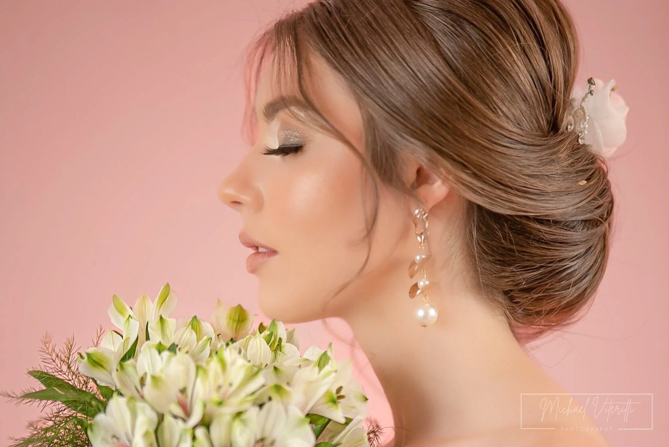 Prima piano di una donna con capelli castani raccolti con accessori e orecchini pendenti, sfondo rosa, in primo piano un bouquet di fiori bianchi con foglie verdi.