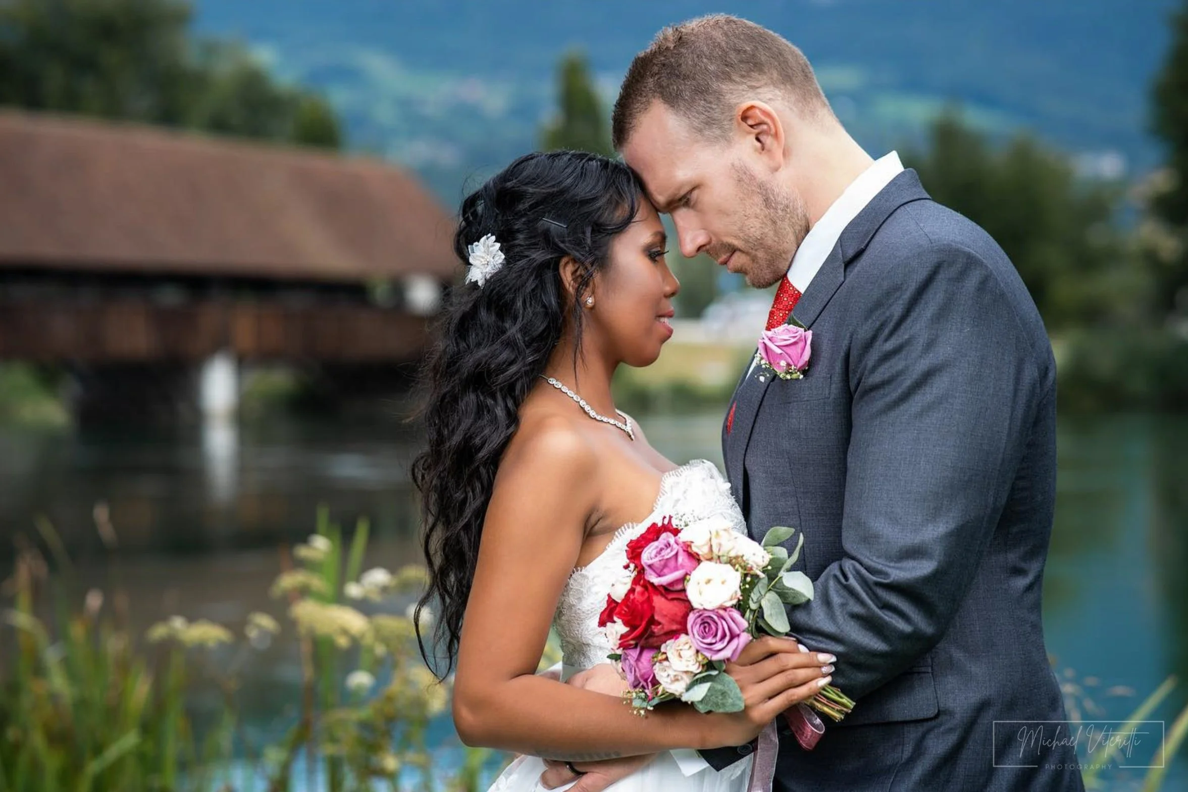 Una coppia di sposi si abbraccia vicino a un lago, con uno sfondo di montagne e vegetazione, celebrando il loro matrimonio outdoor.