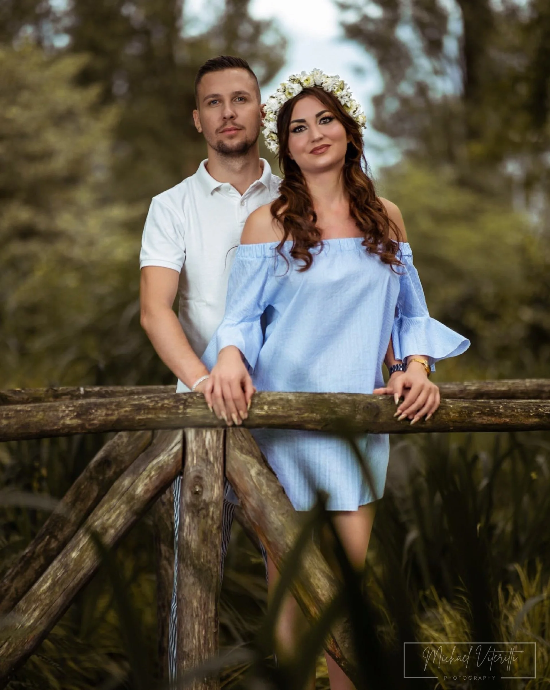 Una coppia di giovani, un uomo e una donna, si tengono per mano davanti a una ringhiera di legno in un ambiente naturale con alberi e foglie verdi. La donna indossa un fiore tra i capelli e un abito blu, mentre l'uomo indossa una camicia bianca.