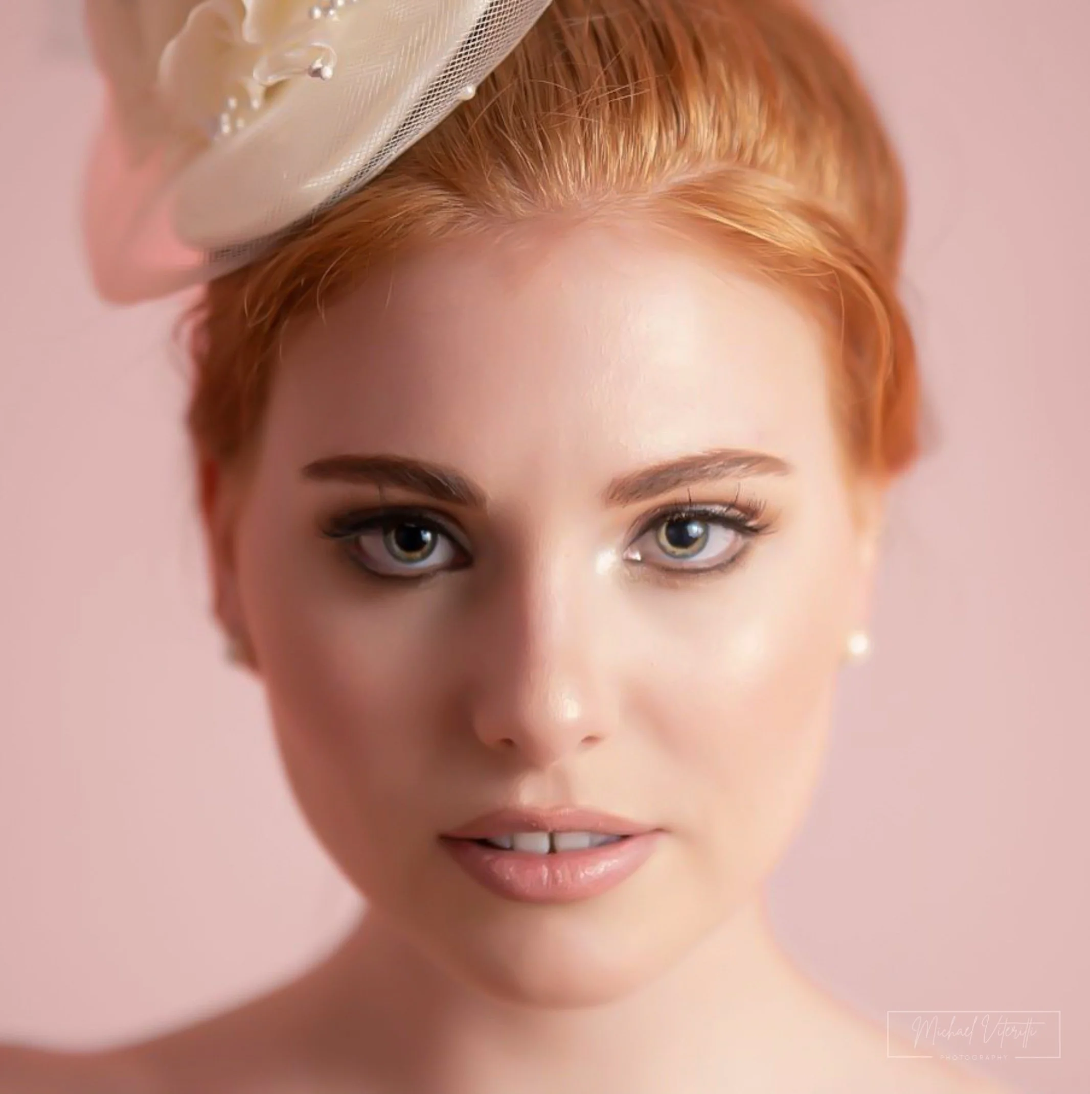 Primo piano di una donna con capelli rossi, occhi chiari e sorriso, sfondo rosa pallido.