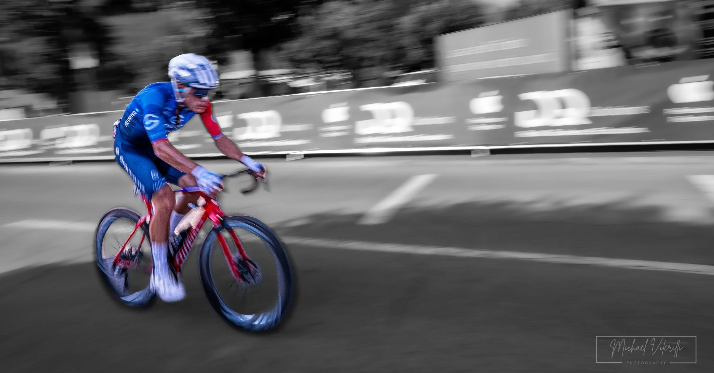 Un ciclista in movimento in un percorso urbano, con sfondo sfocato in bianco e nero, indossa un casco e una maglietta blu