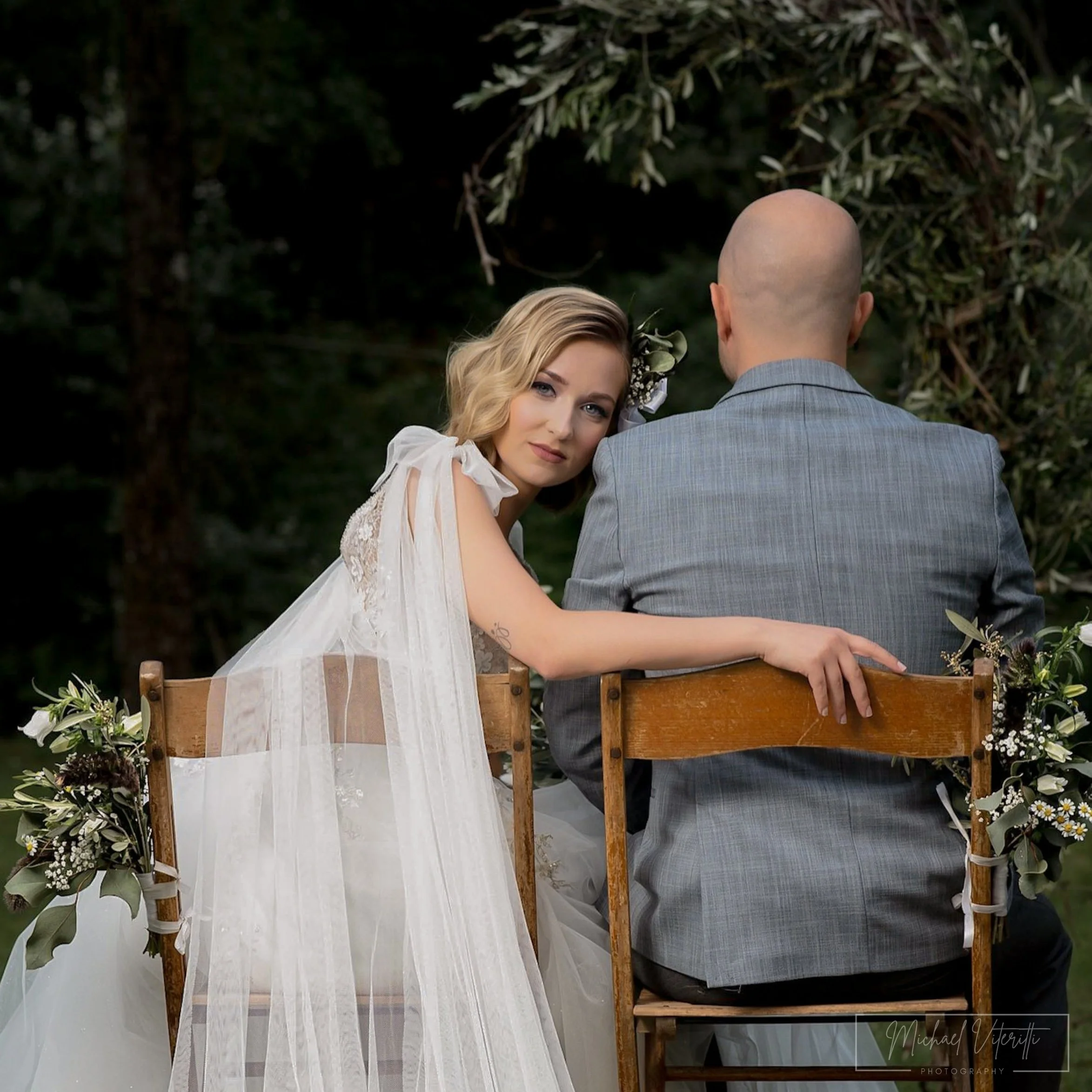 Donna con capelli biondi e trucco naturale, posa con un uomo calvo vestito in grigio, in un luogo all'aperto con alberi e fiori, durante un matrimonio o evento speciale.