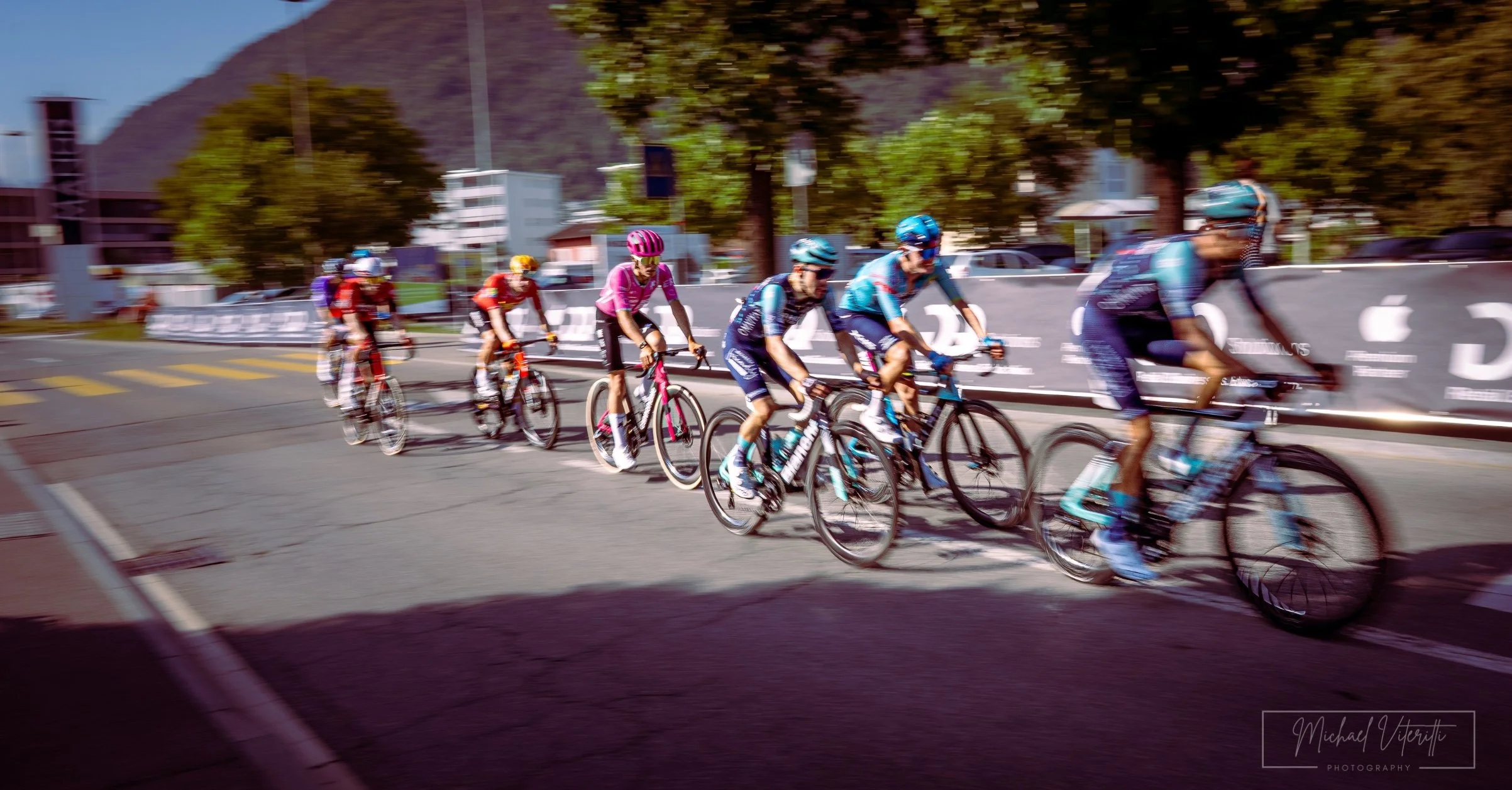 Gruppo di ciclisti che pedala lungo una strada, con alberi e edifici sullo sfondo, durante il giorno.