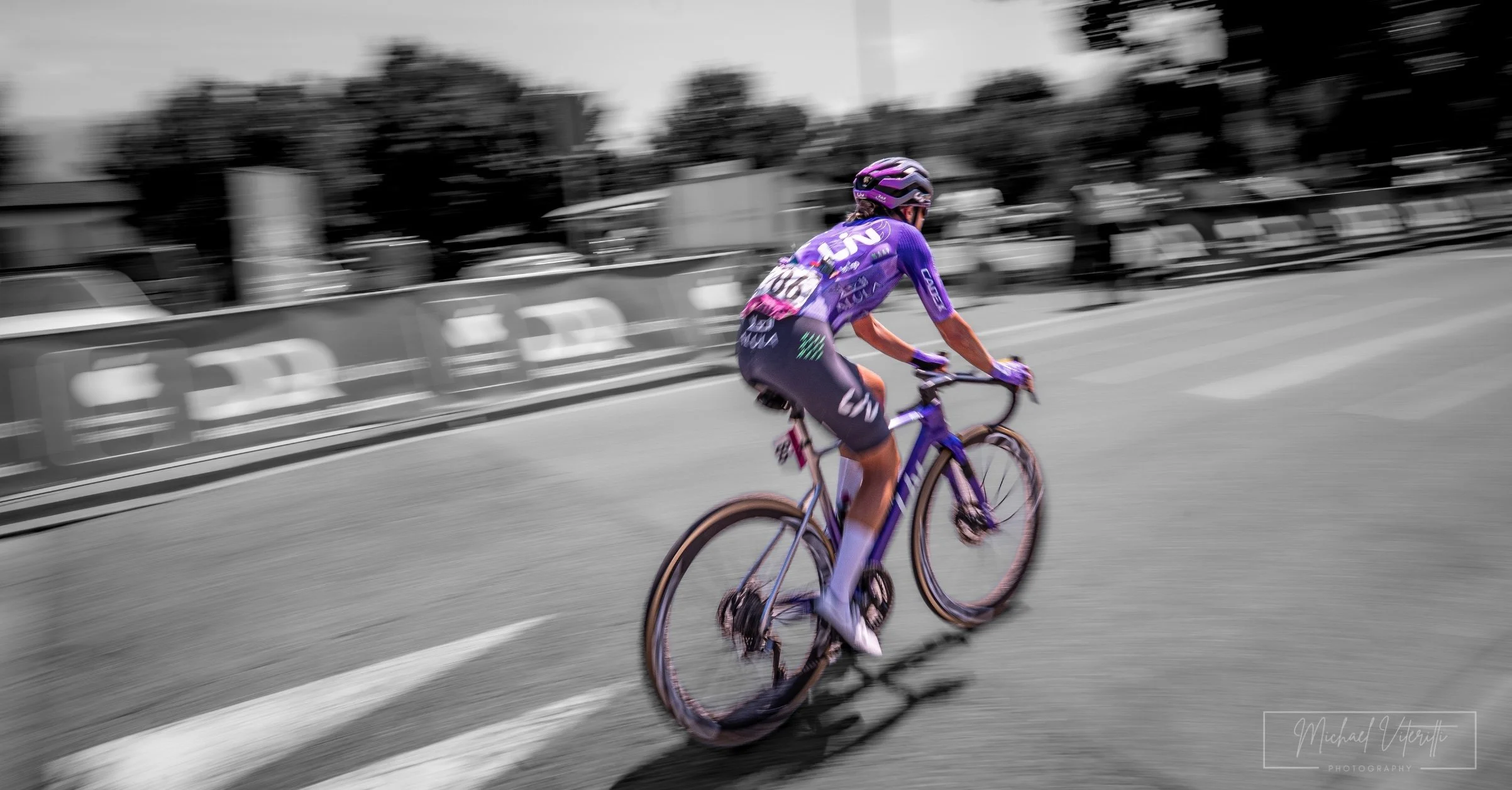 Un ciclista, vestito con un completo viola e casco viola, pedala lungo una strada. L'immagine è in bianco e nero con il ciclista a colori.