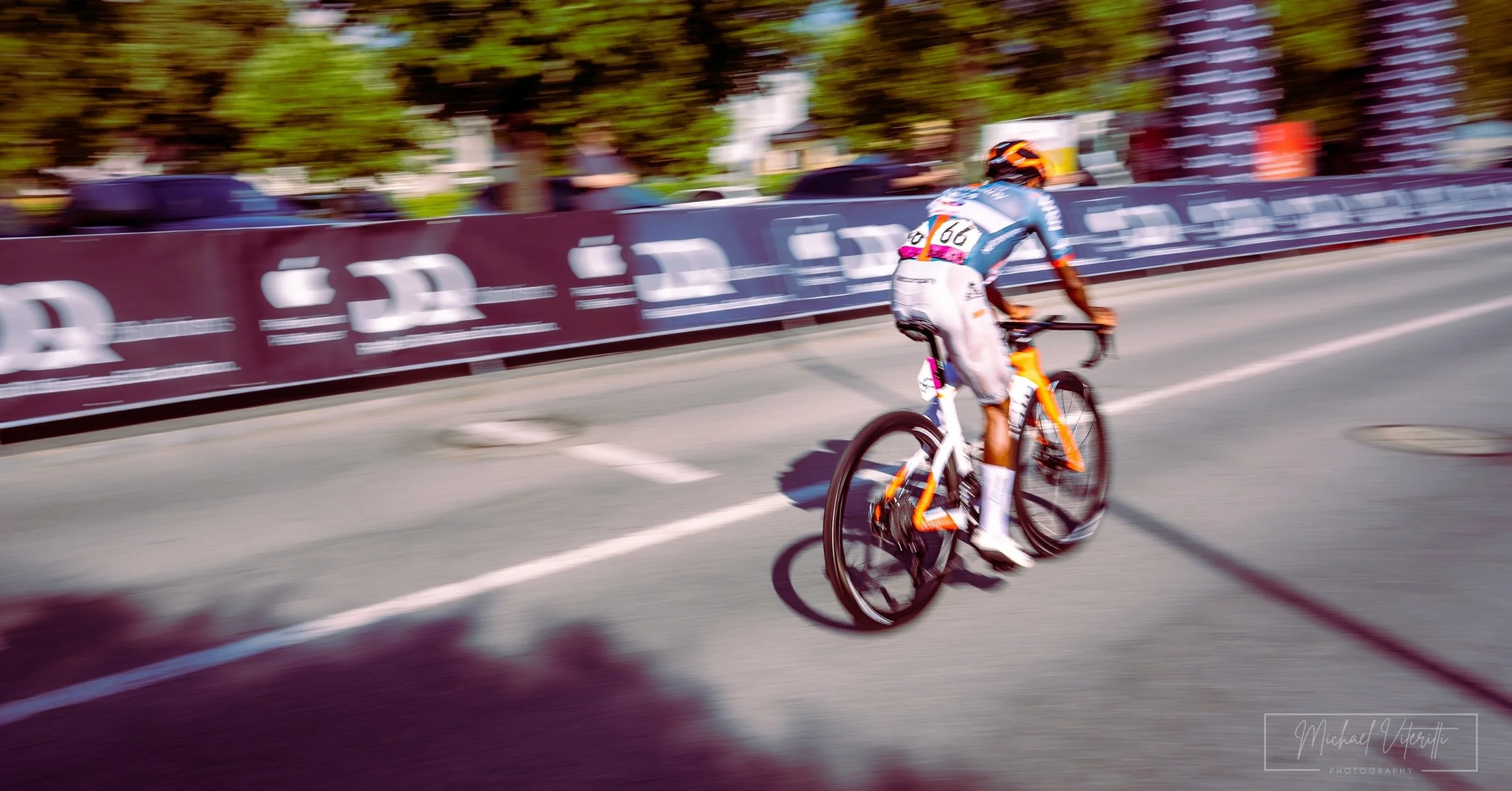 Una persona che partecipa a una gara di ciclismo su una bicicletta da corsa, che attraversa un percorso con barriere pubblicitarie lungo la strada, con alberi e alcune persone sullo sfondo.