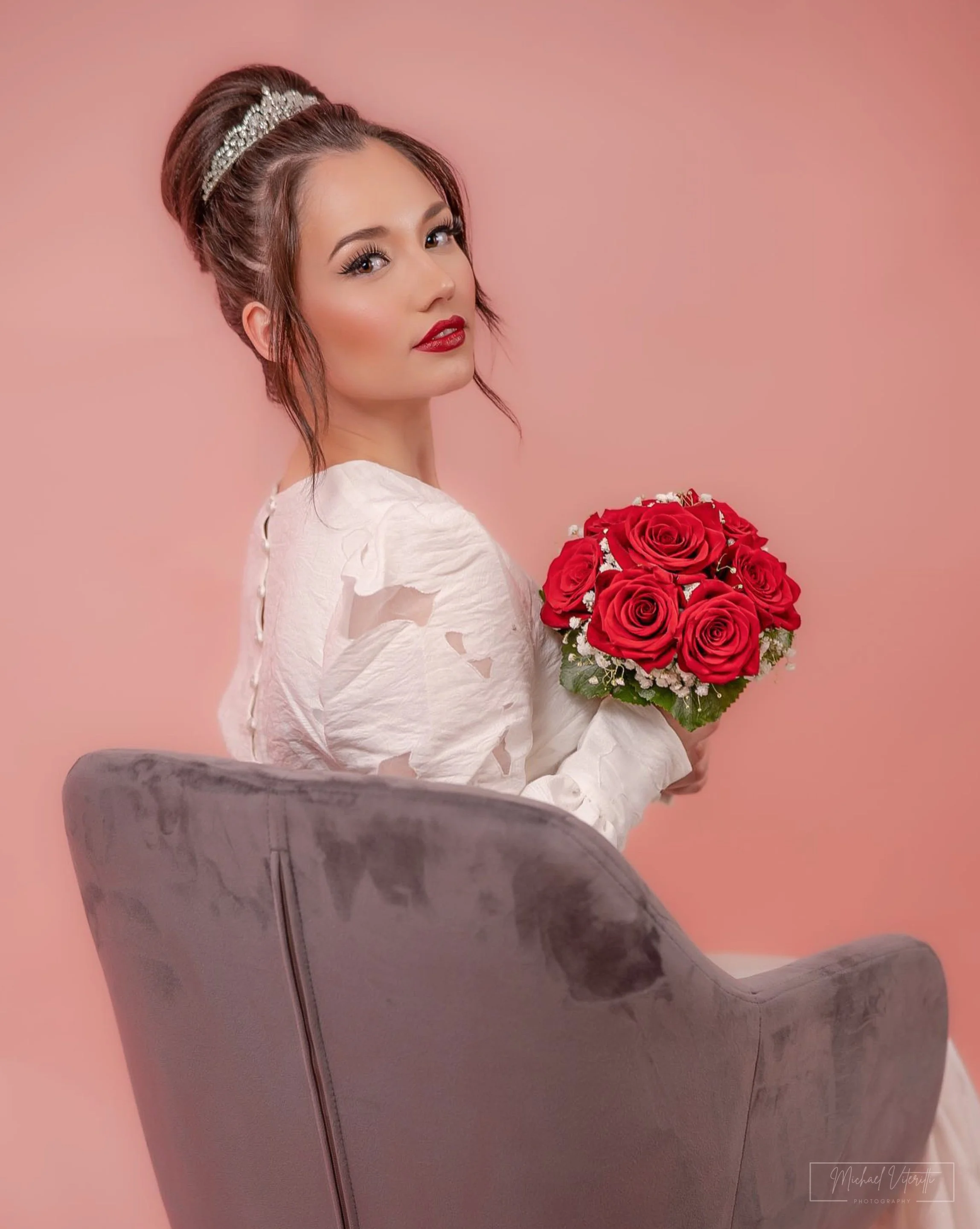 Una donna con trucco elegante, acconciatura raccolta decorata con un cerchietto dorato, che tiene in mano un mazzo di rose rosse e bianco, di fronte a uno sfondo rosa pallido.