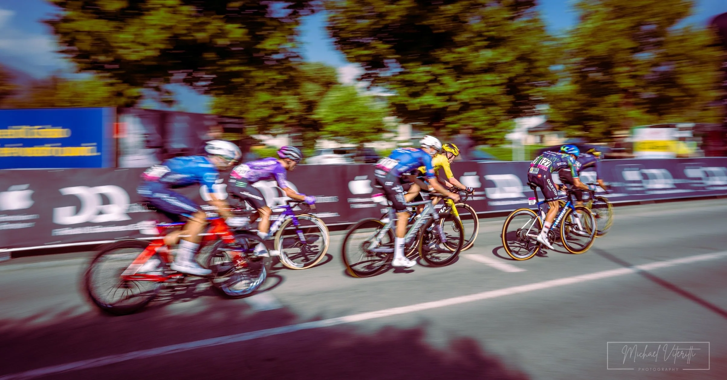 Gruppo di ciclisti che partecipano a una gara, che pedala velocemente lungo una strada con alberi e pubblicità lungo il lato.