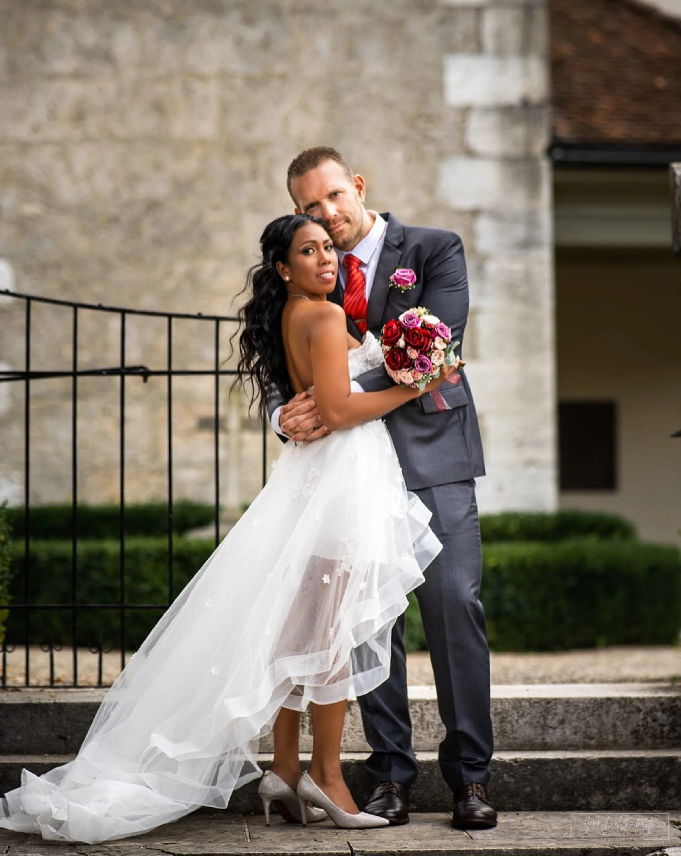 Una coppia di sposi in abiti da matrimonio davanti a un muro di pietra, con il braccio attorno l'uno all'altra, la sposa tiene un bouquet di fiori rosa e rosso, entrambi sorridenti.