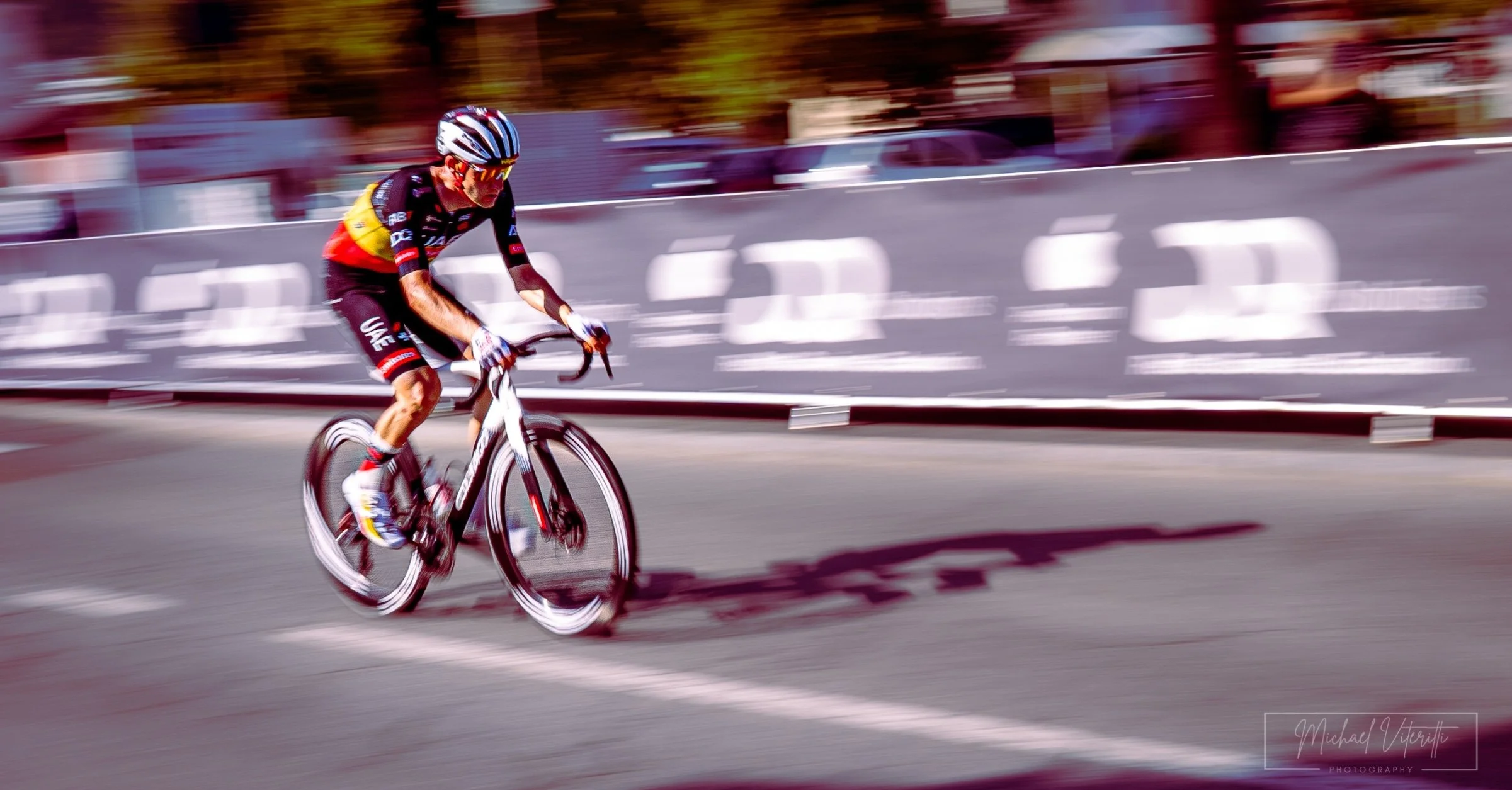 Ciclista in corsa su una strada con sfondo sfocato, indossa casco e abbigliamento sportivo con colori nero, rosso e giallo, pedala su una bicicletta da corsa.