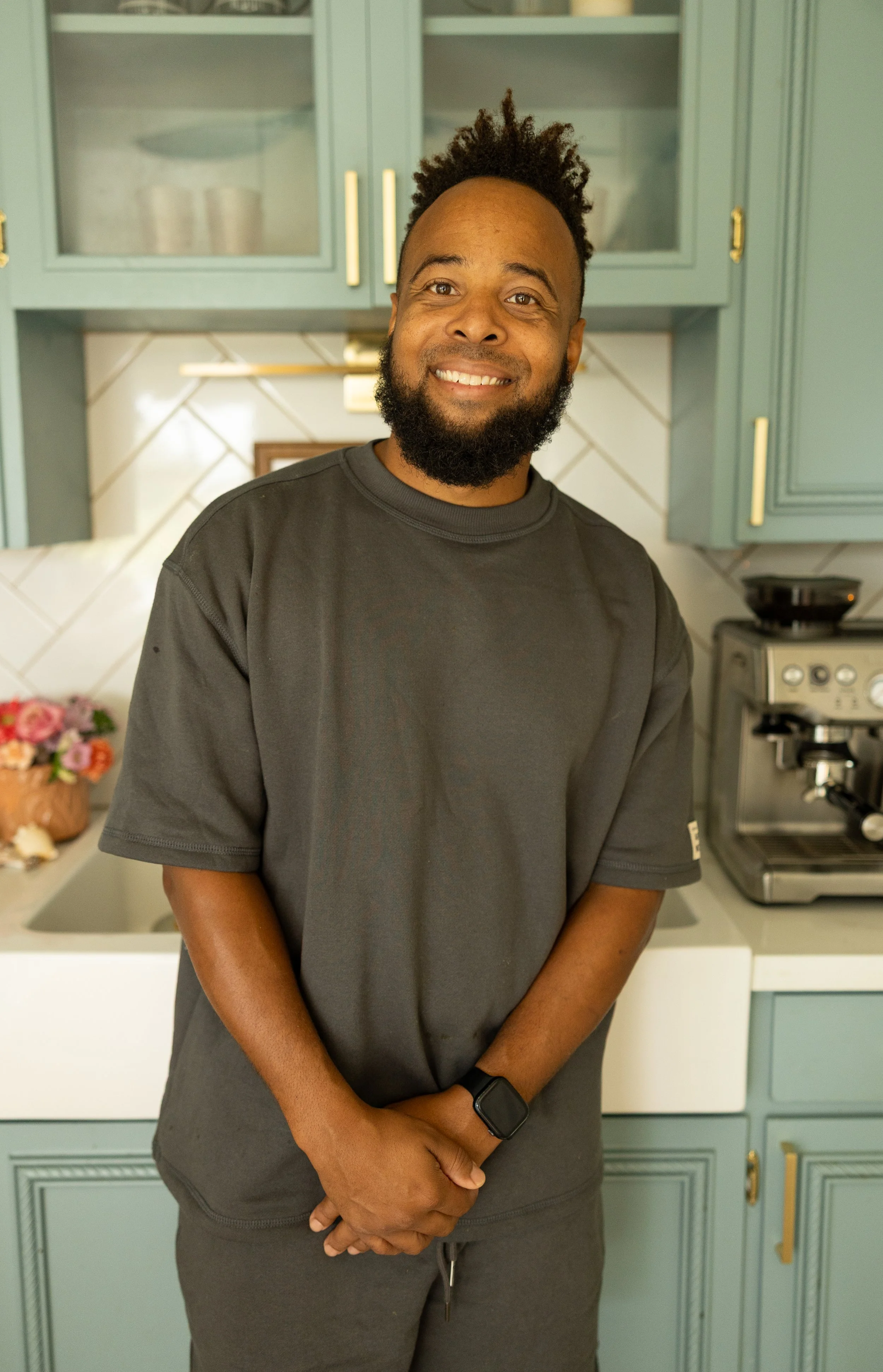A smiling man with a beard and curly hair standing in a kitchen with light blue cabinets, a stove, and a flower bouquet on the counter.