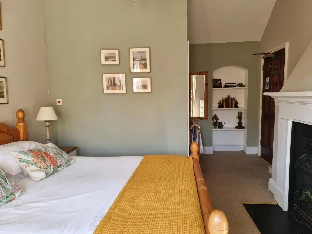 A cozy bedroom with a wooden bed, white bedding, floral pillows, a yellow blanket, a bedside lamp, and a small wall mirror. There is a white fireplace and built-in bookshelf in the background.
