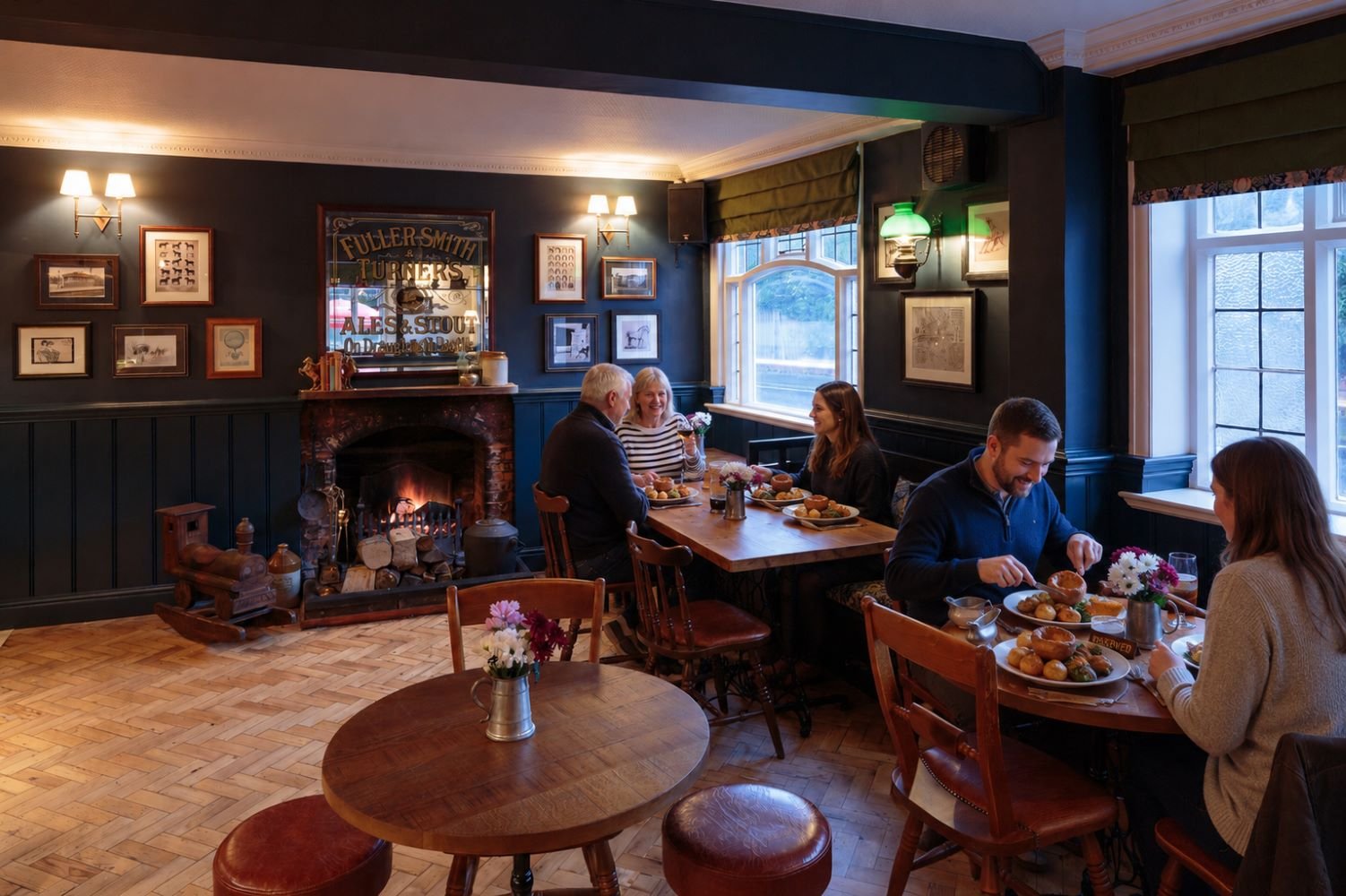 People sitting at two tables enjoying a meal in a cozy, traditional restaurant with a fireplace, dark blue walls, and framed pictures on the wall.