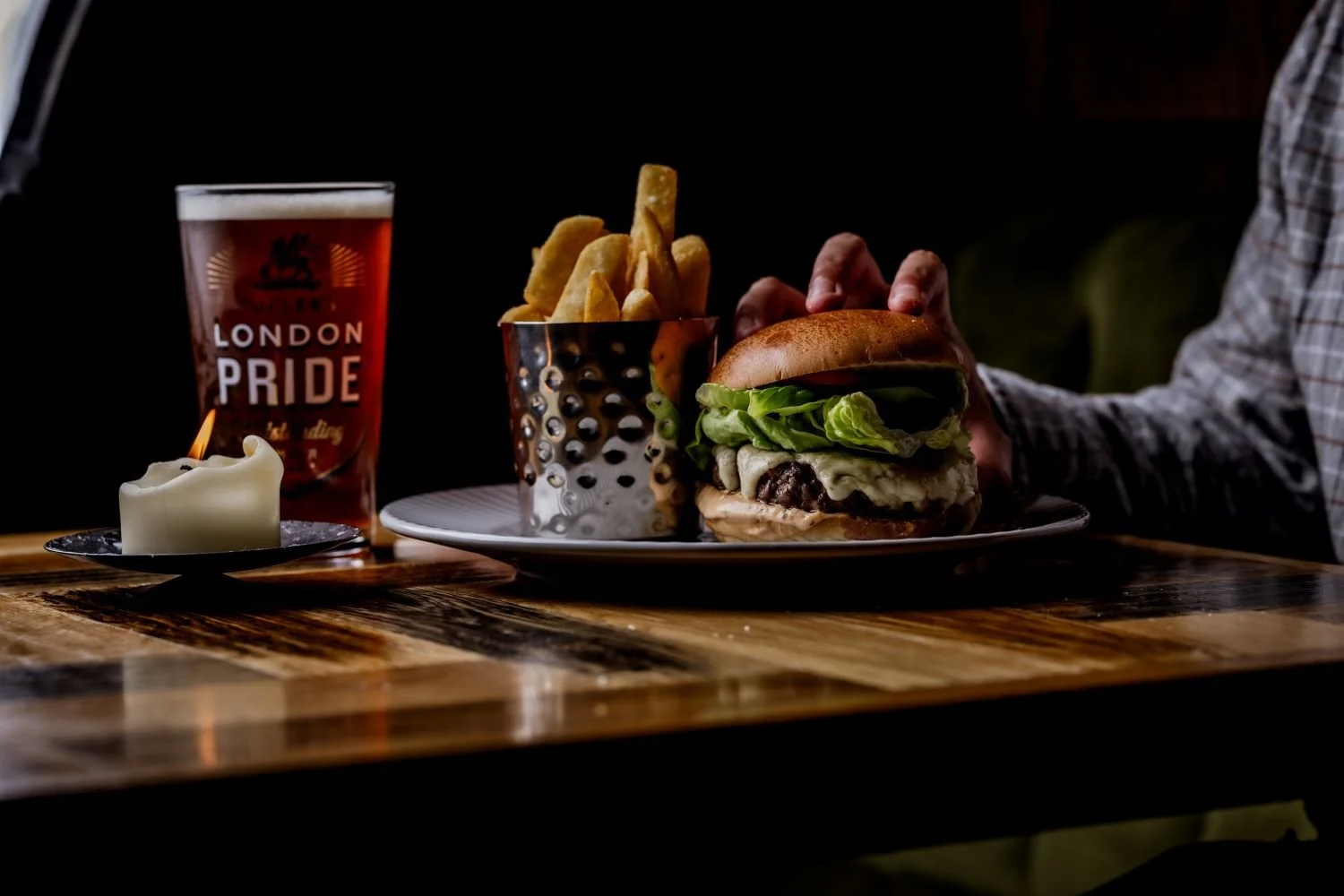 A burger with lettuce, cheese, and meat in a bun, a side of potato fries in a metallic container, a glass of beer with a London Pride label, and a lit candle on a wooden table.