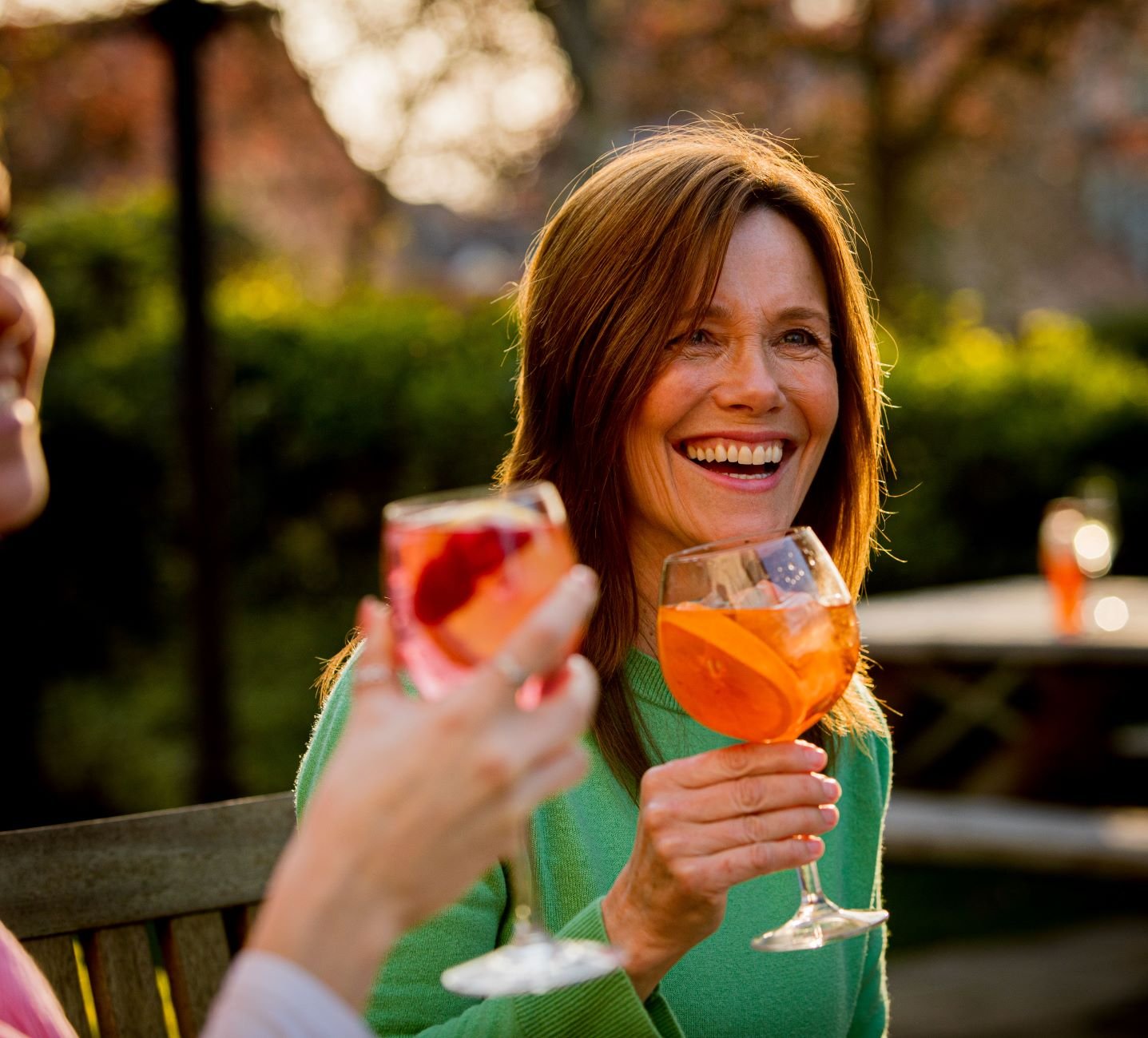 A woman with light brown hair, smiling and holding a large glass of orange-colored drink with orange slices, enjoying an outdoor gathering during sunset.