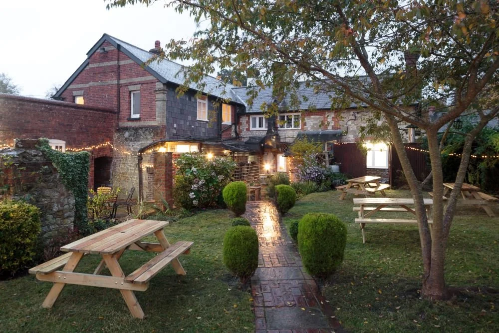 A cozy backyard patio at dusk with string lights, picnic tables, a brick walkway, and a brick house with a stone and brick facade.