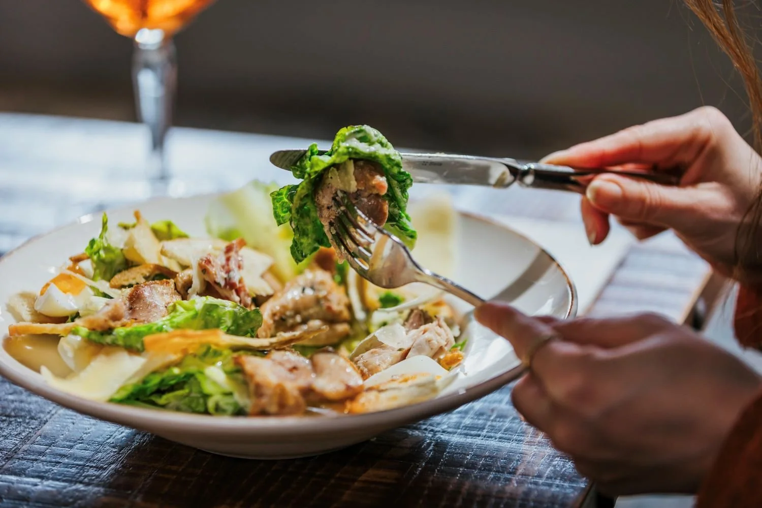 Person holding a fork with a bite of salad above a white bowl of mixed salad on a wooden table.