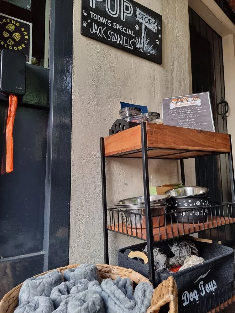 A signboard advertising daily specials and Jack Daniels at a pet-friendly cafe. A shelf with pet bowls and a basket with gray pet blankets or toys are visible.