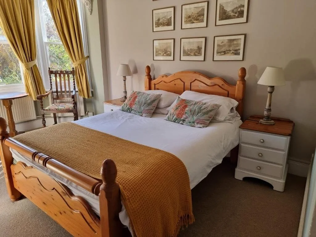 Bedroom with a wooden bed frame, white bedding, floral pillows, yellow blanket, two white bedside tables with lamps, a chair by the window, and framed artwork on the wall.