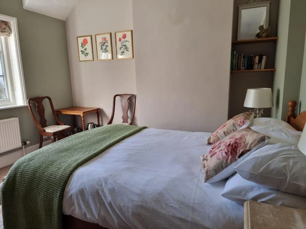 Bedroom with a double bed, green blanket, floral pillows, bedside lamps, bookshelf, mirror, desk, chairs, window, and framed floral art on the wall.