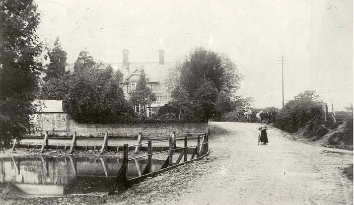 An old black-and-white photograph of a rural dirt road with a woman walking away from the camera, houses with large trees in the background, and a small pond separated by a wooden fence.
