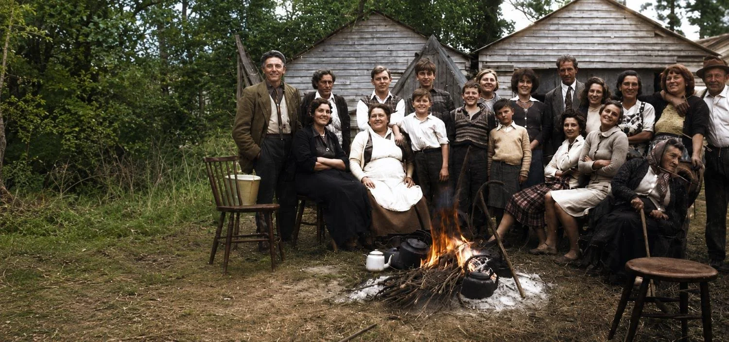A group of people, including men, women, and children, gathered outdoors around a campfire with teapots and logs, in front of wooden structures, with trees in the background.