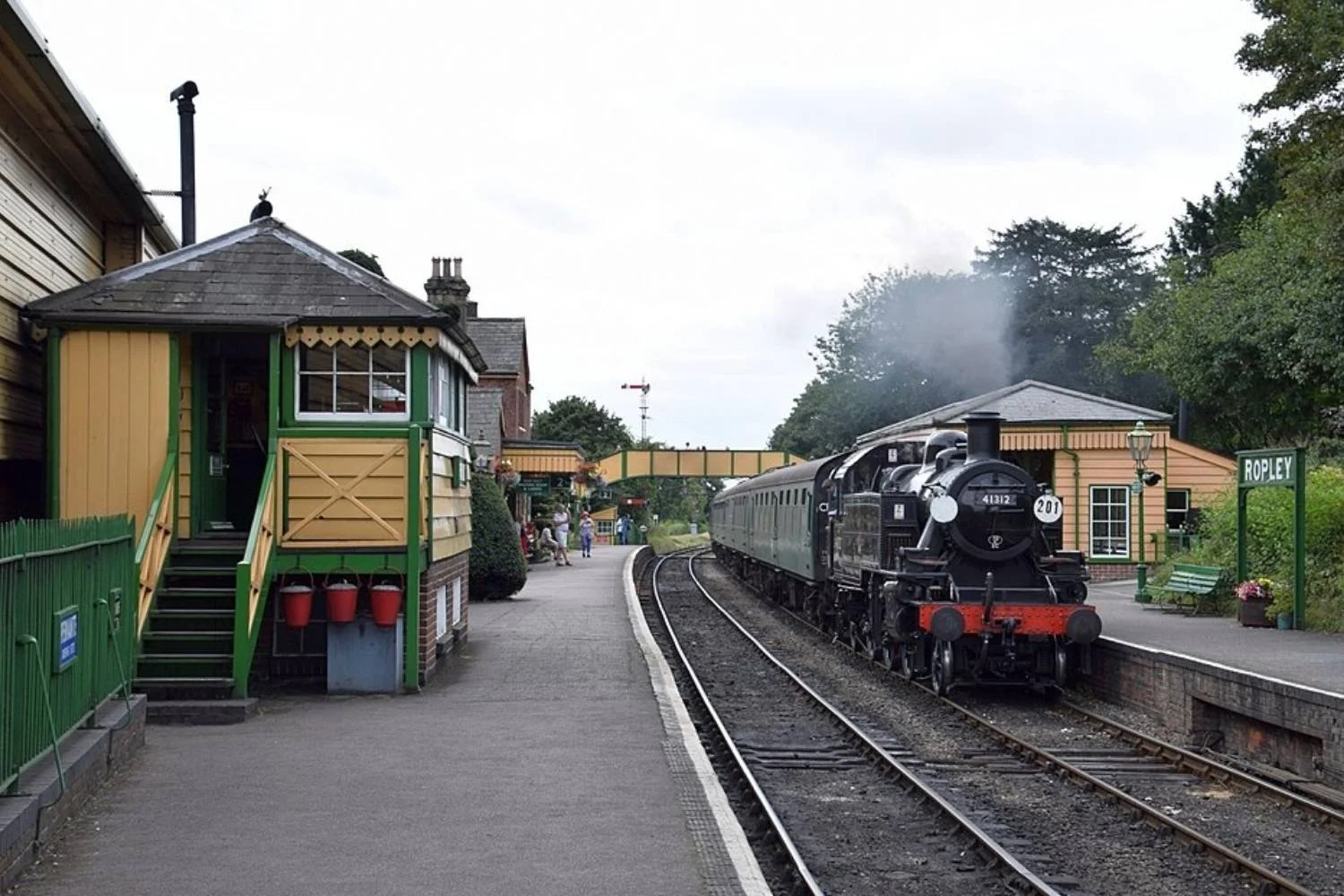 Vintage steam train stopping at a small train station named Ropley, with passengers on the platform and historical buildings nearby.