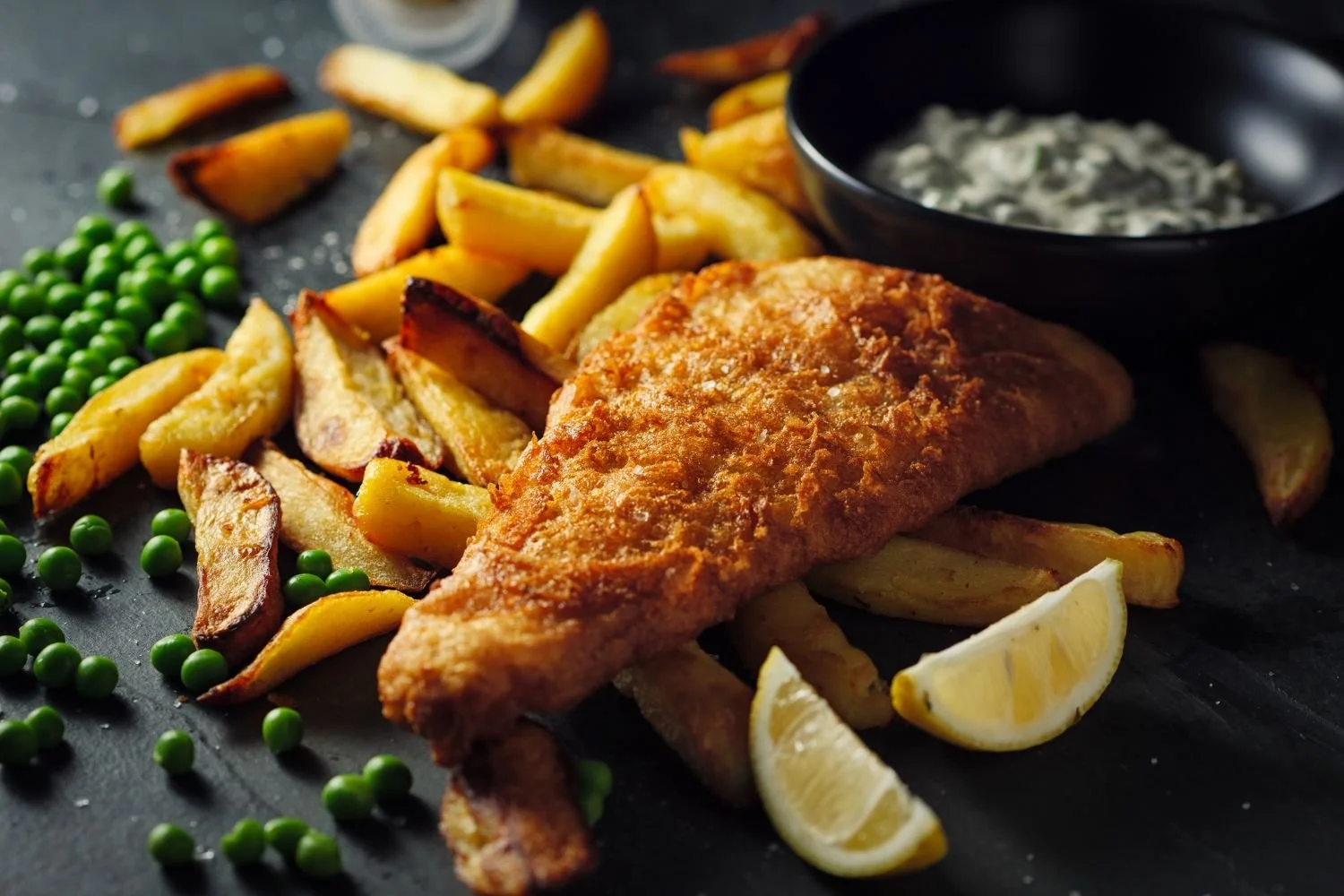 Fried fish fillet with French fries, green peas, lemon wedges, and tartar sauce on a black plate.