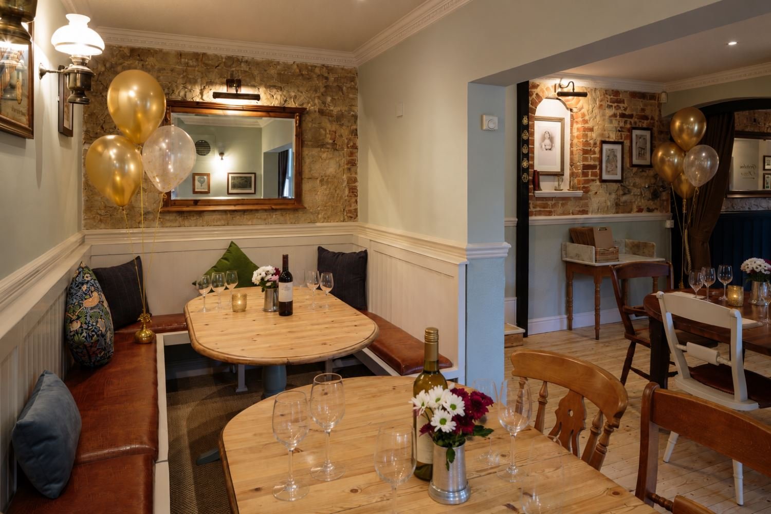 Cozy restaurant with wooden tables, balloons, wine bottles, and decorative pillows, featuring exposed brick walls and framed art.