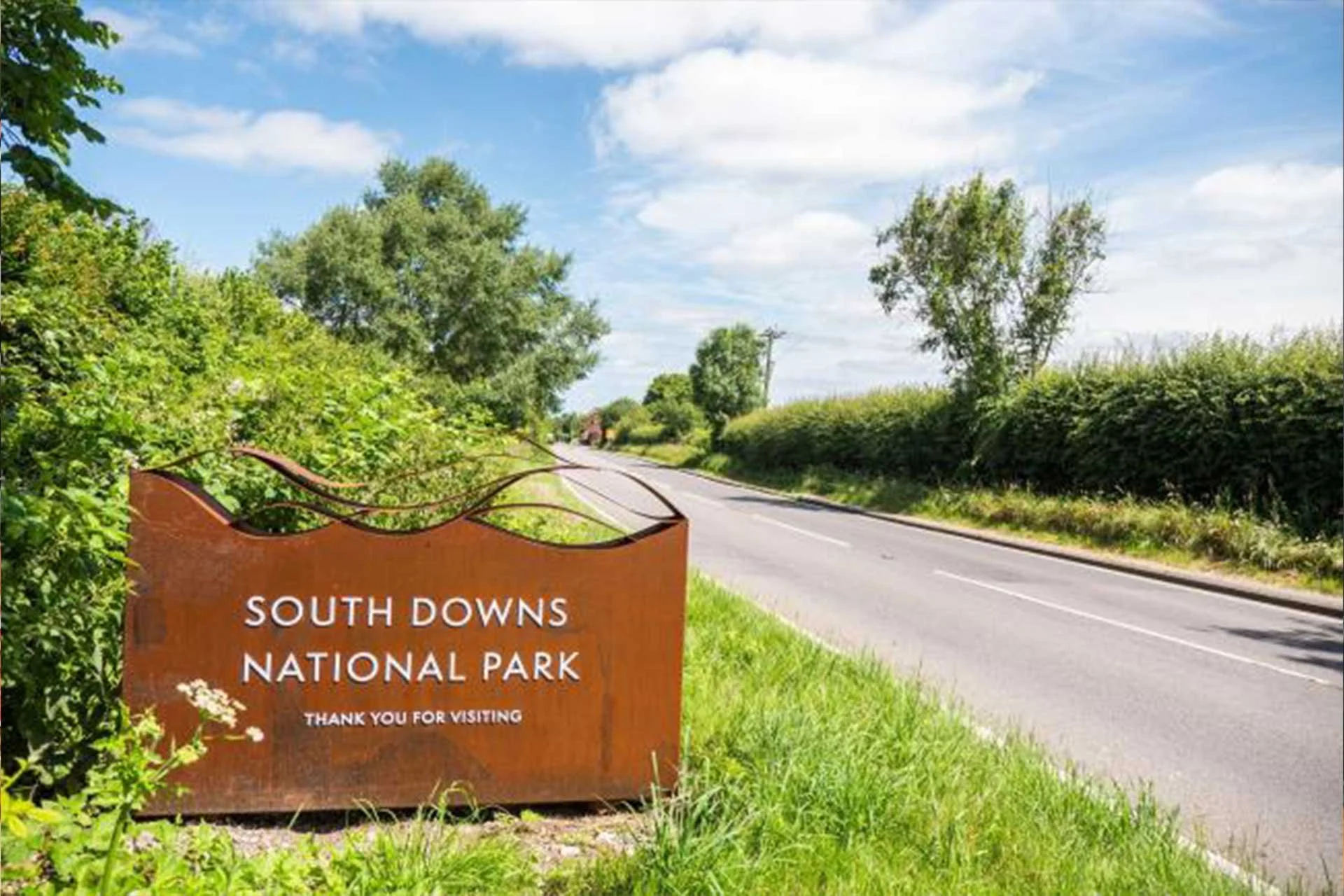 Rust-colored sign that reads 'South Downs National Park, Thank you for visiting' on the roadside with green bushes and trees, a paved road, blue sky, and white clouds in the background.