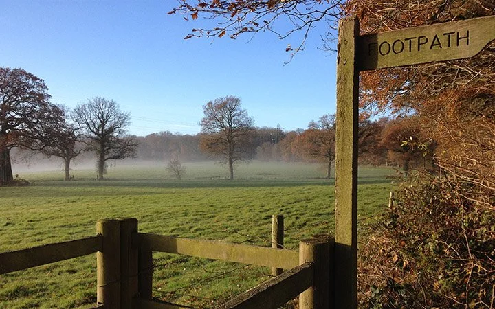 A rural footpath sign next to a wooden fence, overlooking a grassy field with trees and low fog in the distance on a clear day.