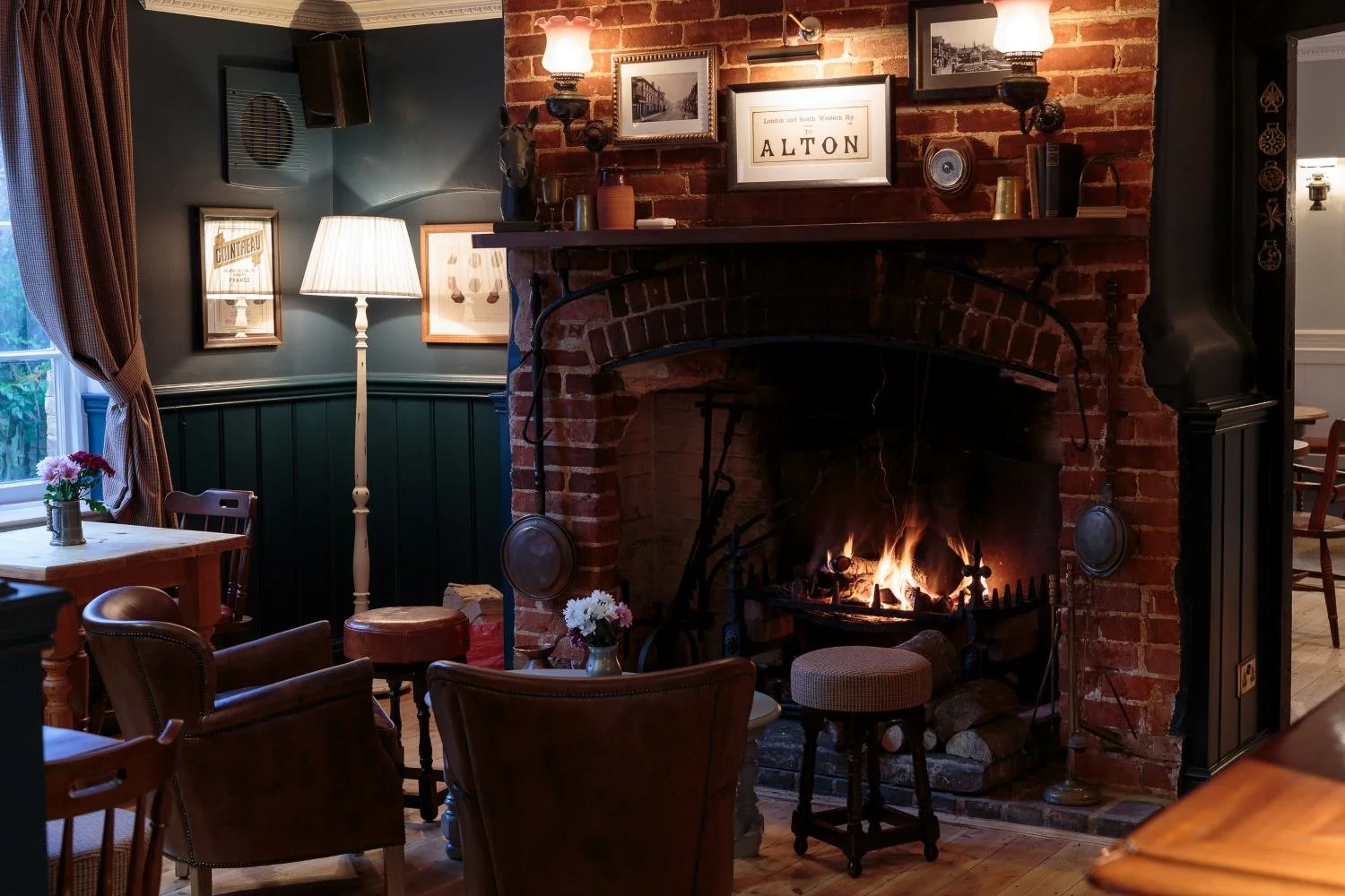 Cozy room with brick fireplace, wood logs burning, surrounded by chairs and a small table with flowers, green paneled walls, framed pictures, and soft lighting.