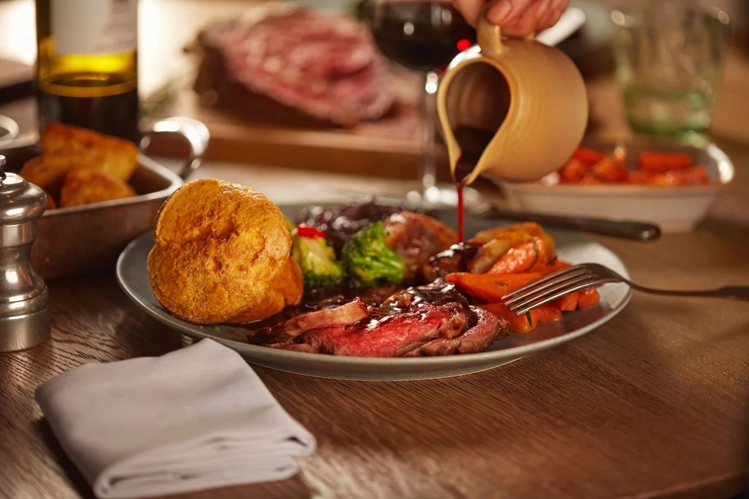 A plate of roast beef, vegetables, and Yorkshire pudding on a wooden table with a fork. In the background, a hand pours gravy over the food, with additional dishes, a glass of wine, and seasoning shakers nearby.
