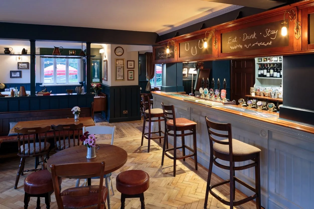 Interior of a cozy pub with wooden furniture, a bar area with beer taps, and a chalkboard sign that says "Eat, Drink, Stay."
