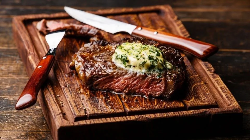 A cooked steak topped with herb butter on a wooden cutting board, accompanied by a steak knife and fork.